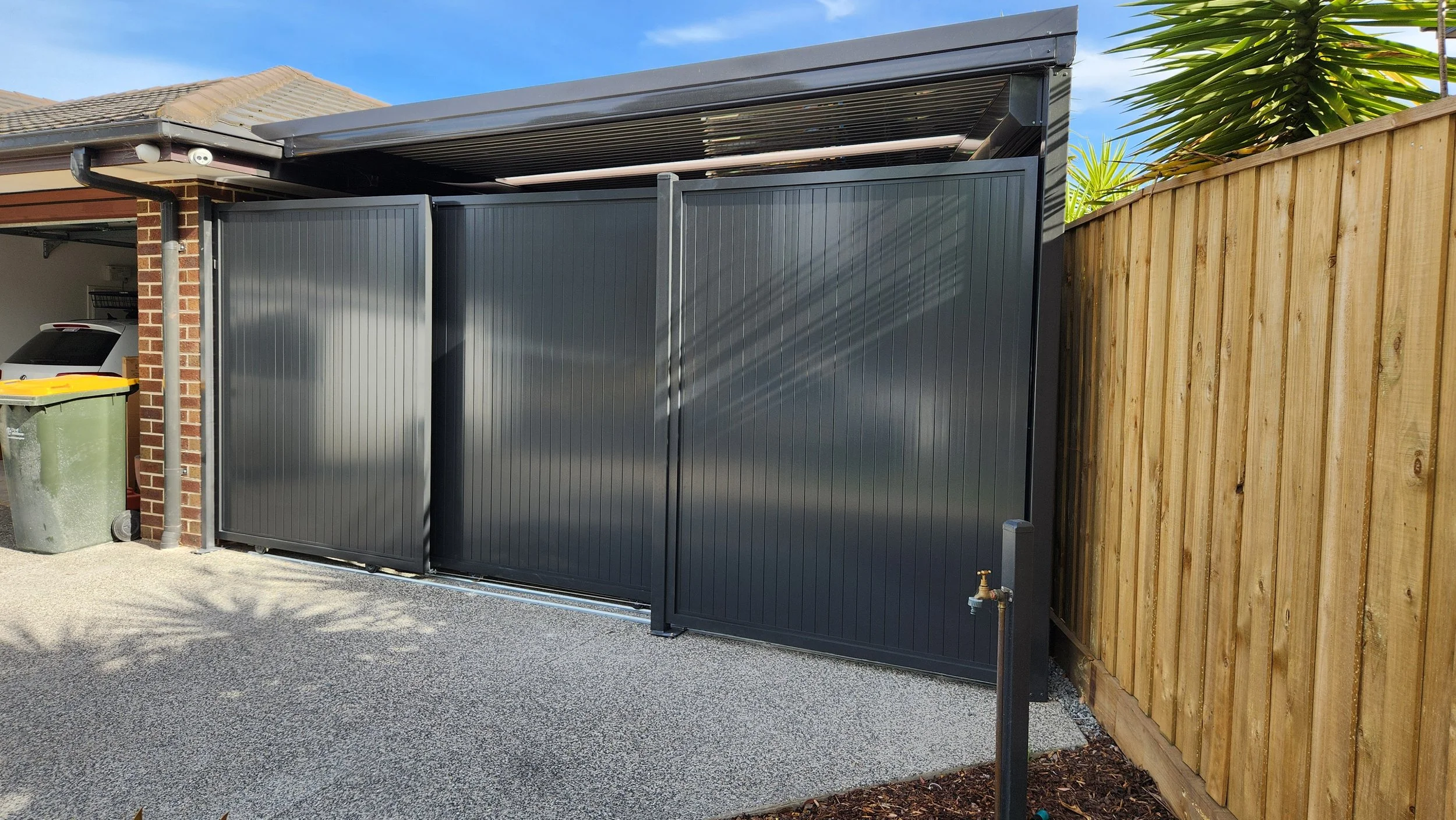 A black metal sliding gate next to a wooden fence in a residential backyard, with a trash bin and part of a garage visible, under a blue sky.