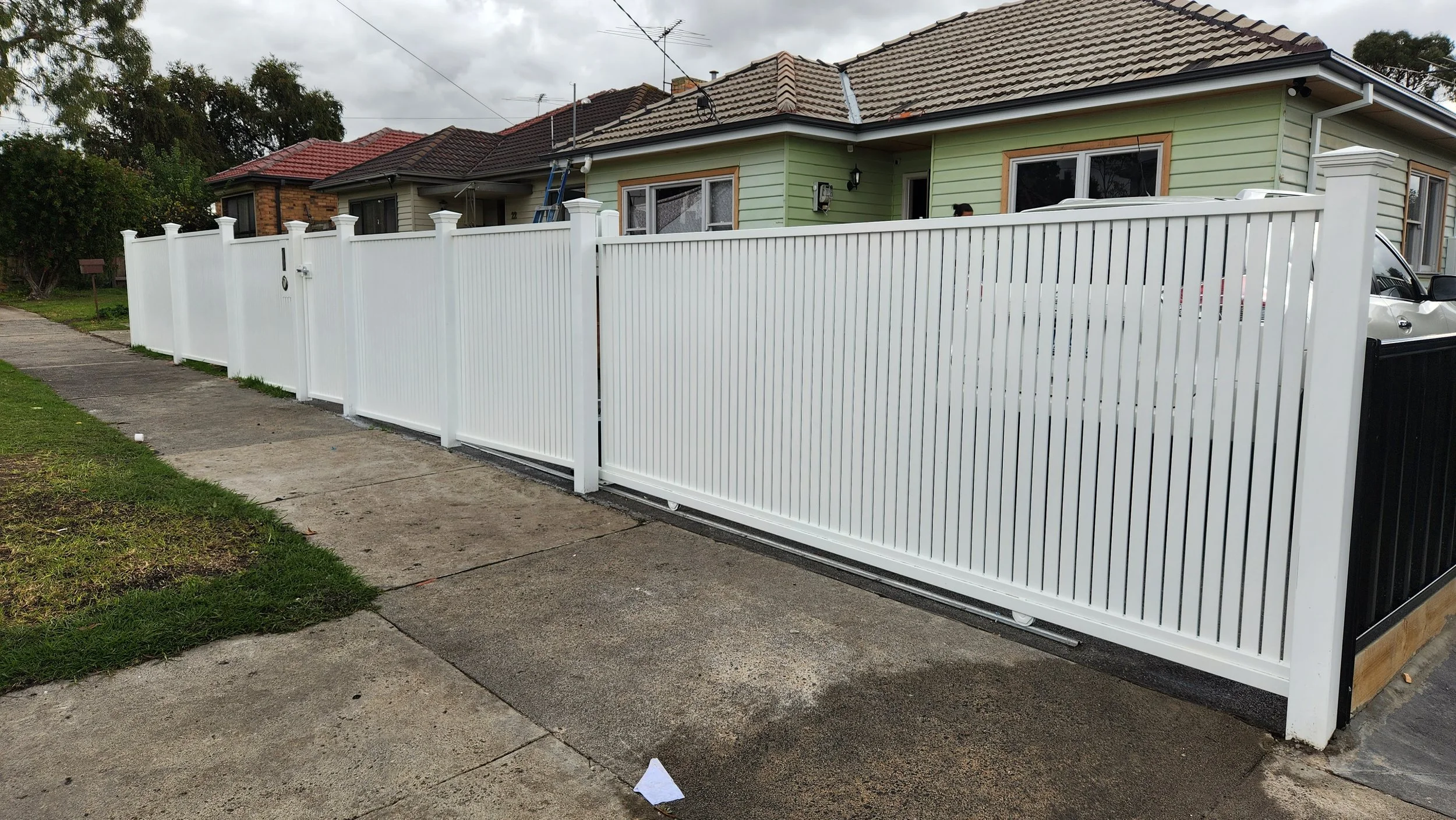 White vinyl fence along a sidewalk in front of a green house with brown trim, with a driveway and parked car. Overcast sky.