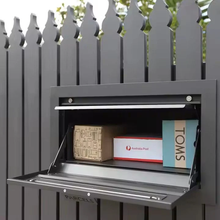A gray mail drop box mounted on a fence, containing three packages with a metal flap open.