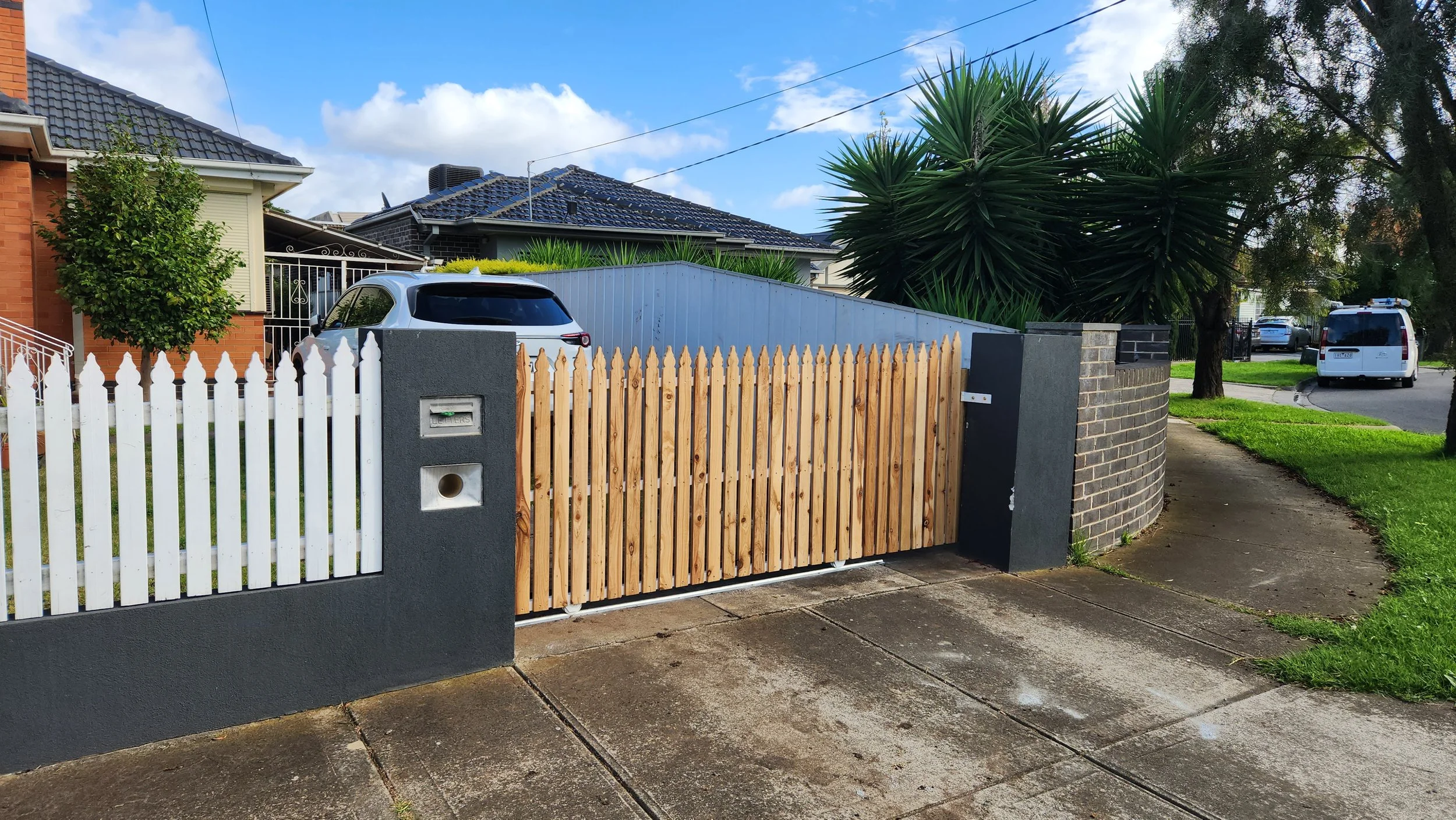 Residential street scene with a white wooden picket fence, a wooden sliding gate, and a gray brick wall. A parked car is visible in the driveway behind the fence. Green trees and houses are in the background under a partly cloudy sky.