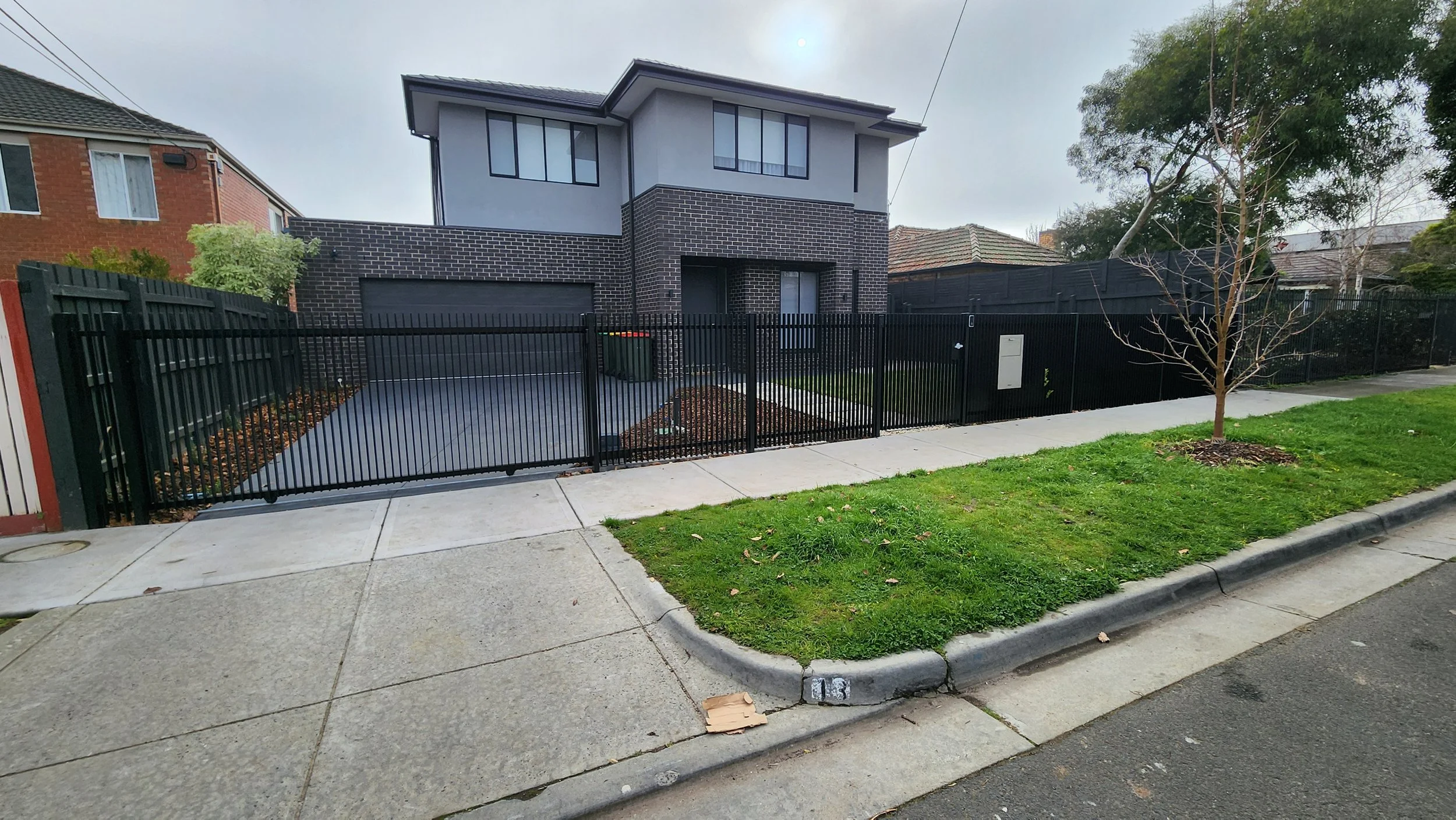 Modern two-story house with black brick and gray siding, fenced yard with a small tree, sidewalk, and green lawn in a residential neighborhood.