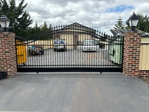 Black metal gate with brick pillars and lamps, open for vehicle entry, leading to a parking area with cars and a garage building.