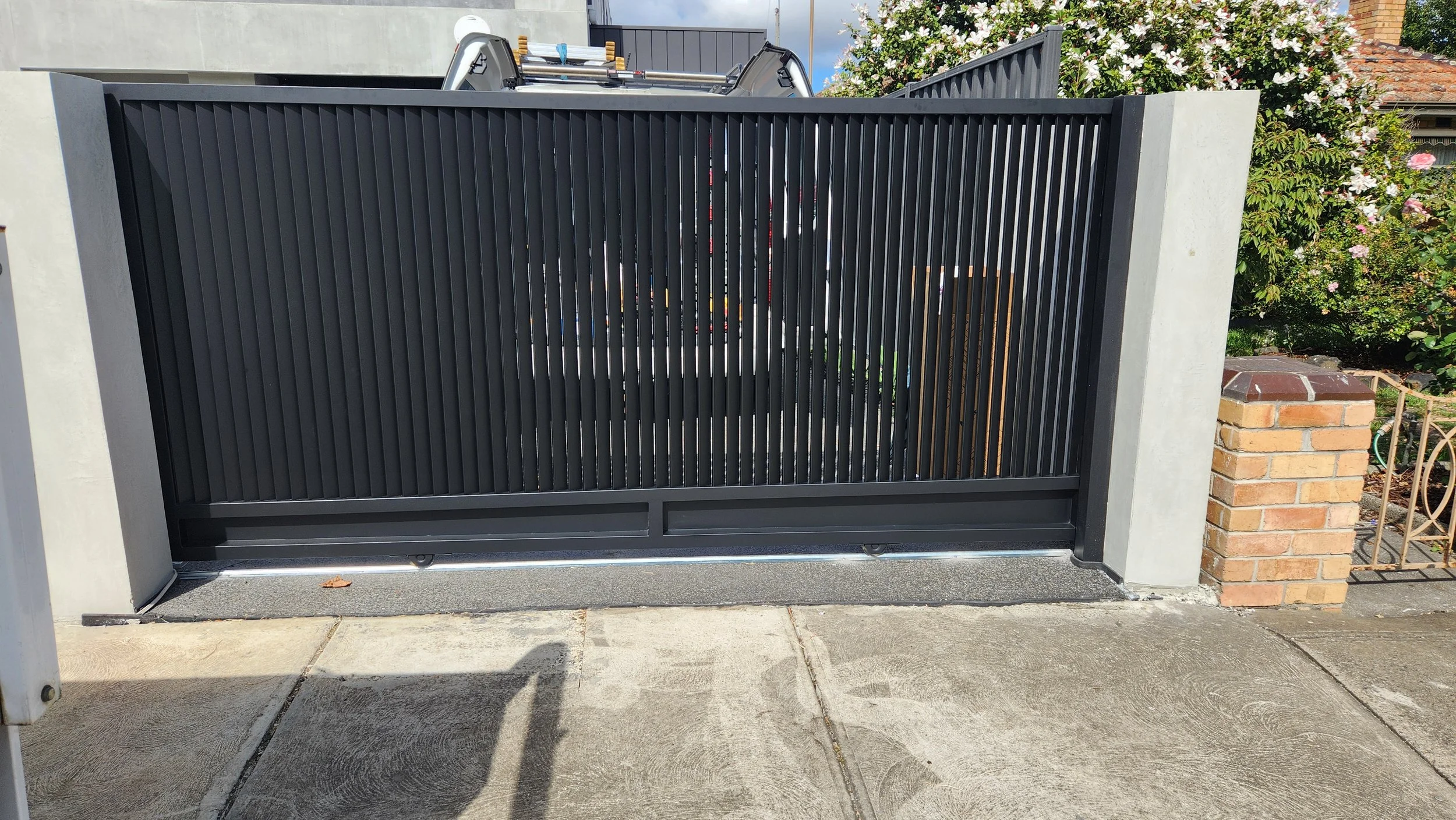 Black metal sliding gate with vertical slats mounted between concrete pillars, with a brick pillar and flowering bushes in the background.