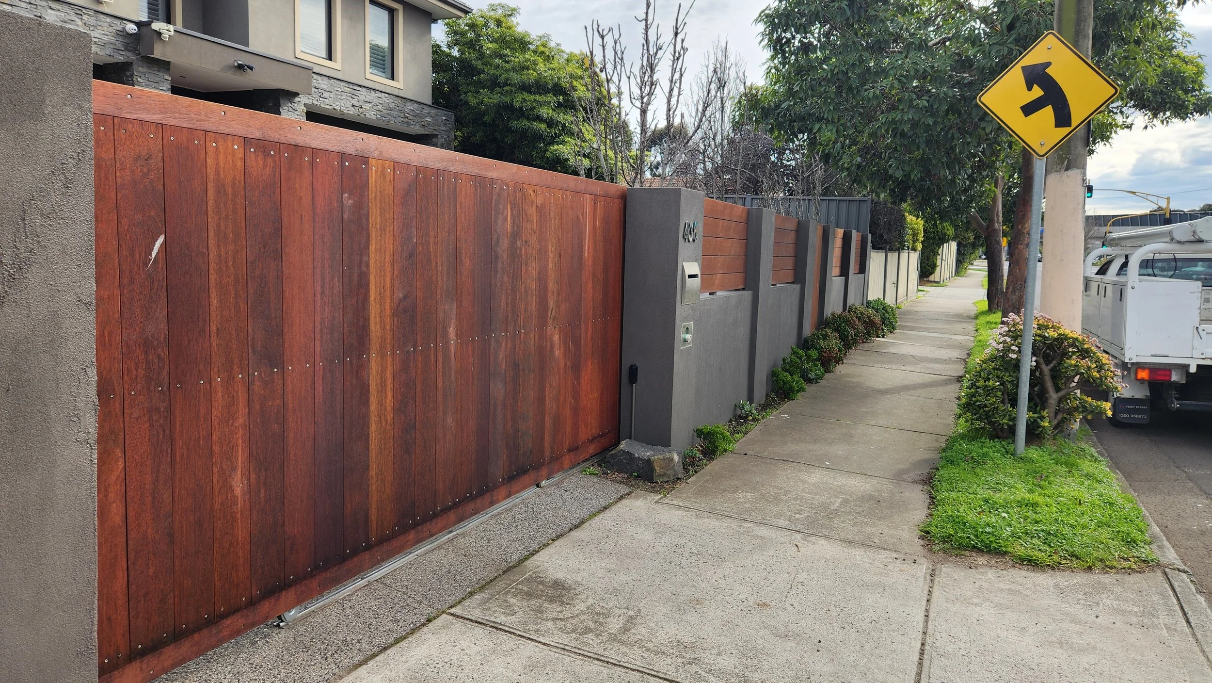 Residential street with a wooden and gray concrete fence, a sidewalk, trees, a yellow right turn traffic sign, and a parked truck.