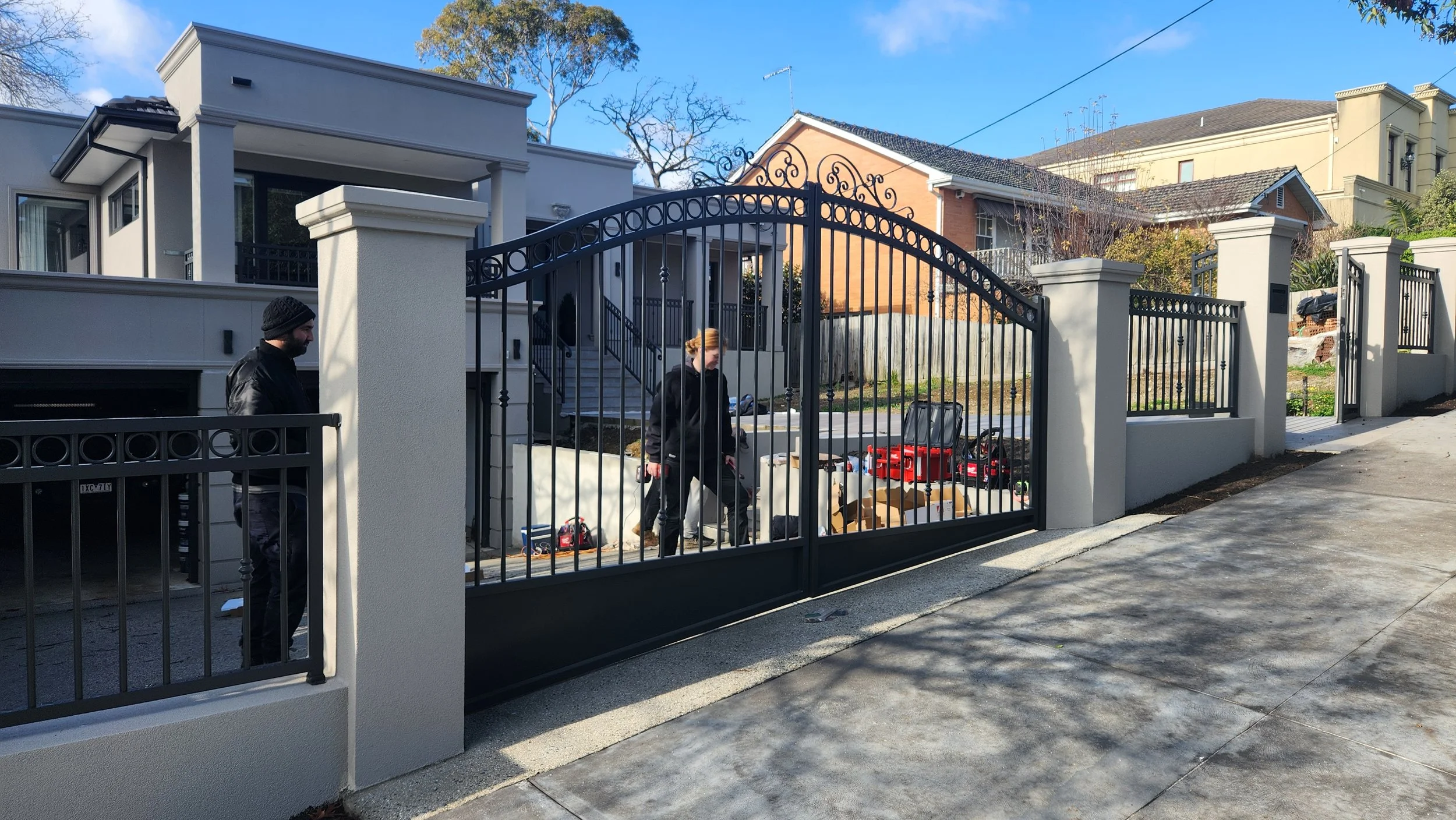 Two people in black jackets and a woman with red hair walking by a black metal gate in front of a house, with some tools and boxes on the ground, on a sunny day.