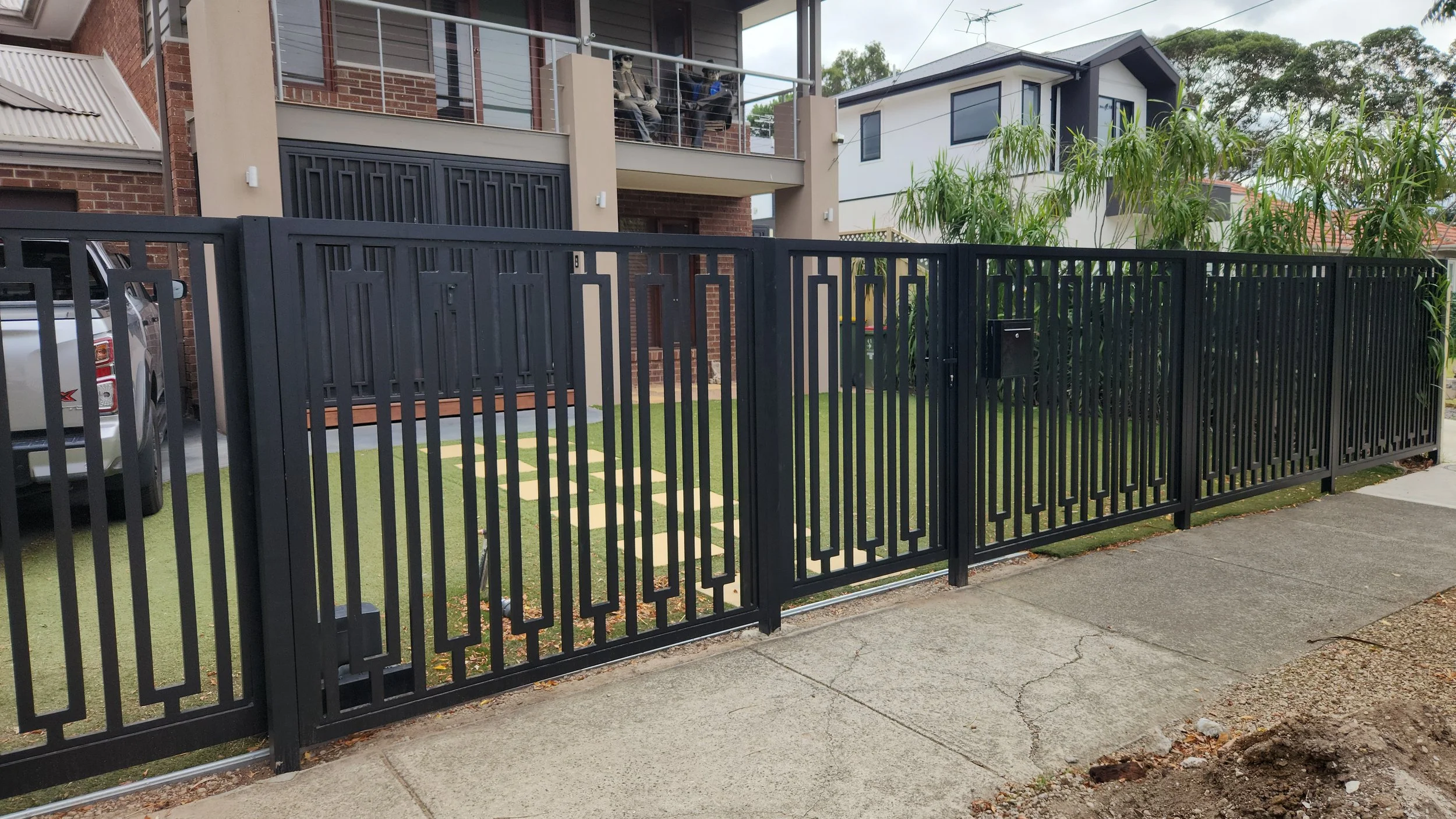 A black metal fence with a gate surrounds a modern two-story house with a small front yard and a sidewalk in front.