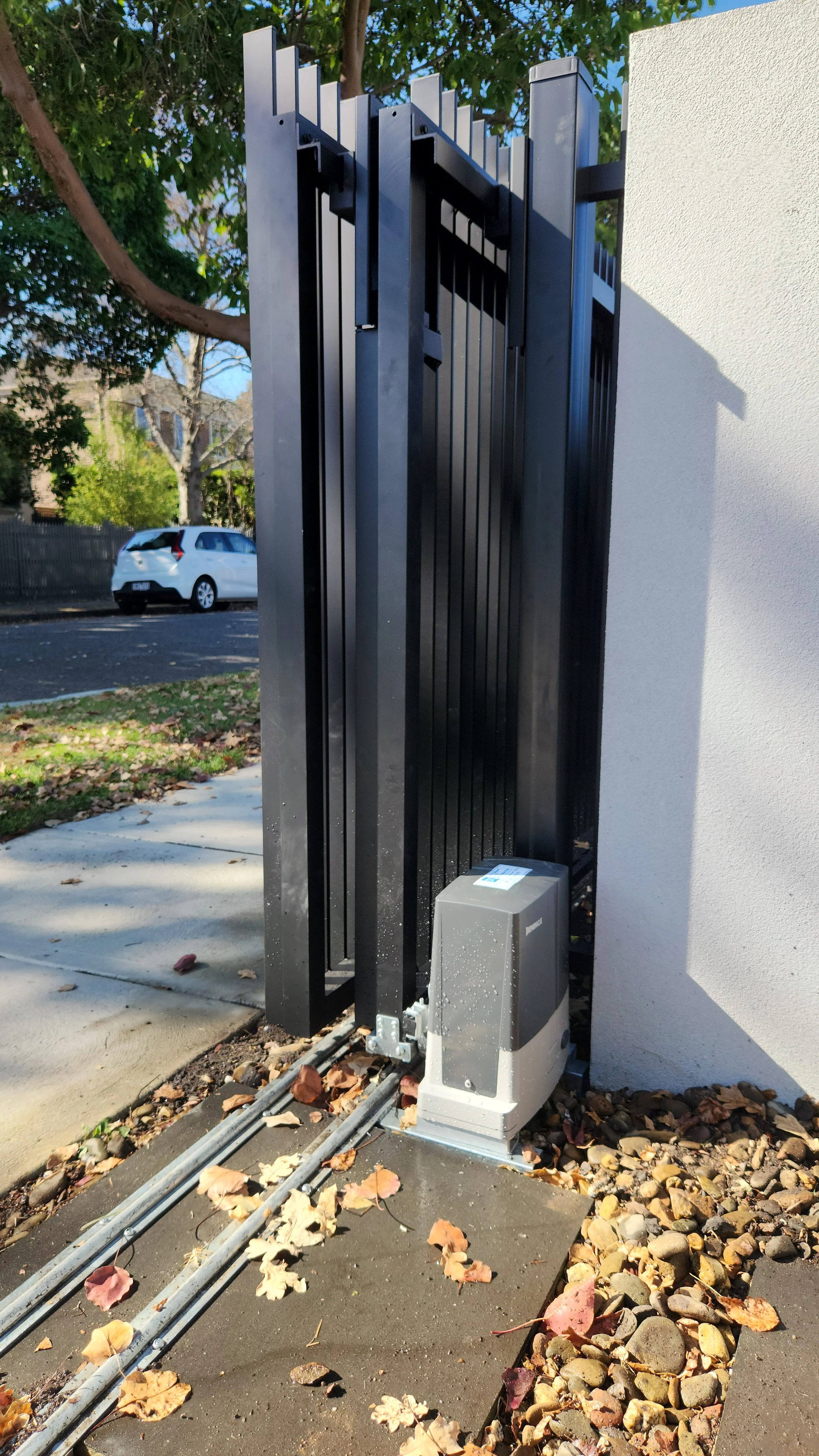 Automated sliding gate installed beside a white wall with gate motor at the bottom, surrounded by fallen leaves and small rocks, and green trees in the background.