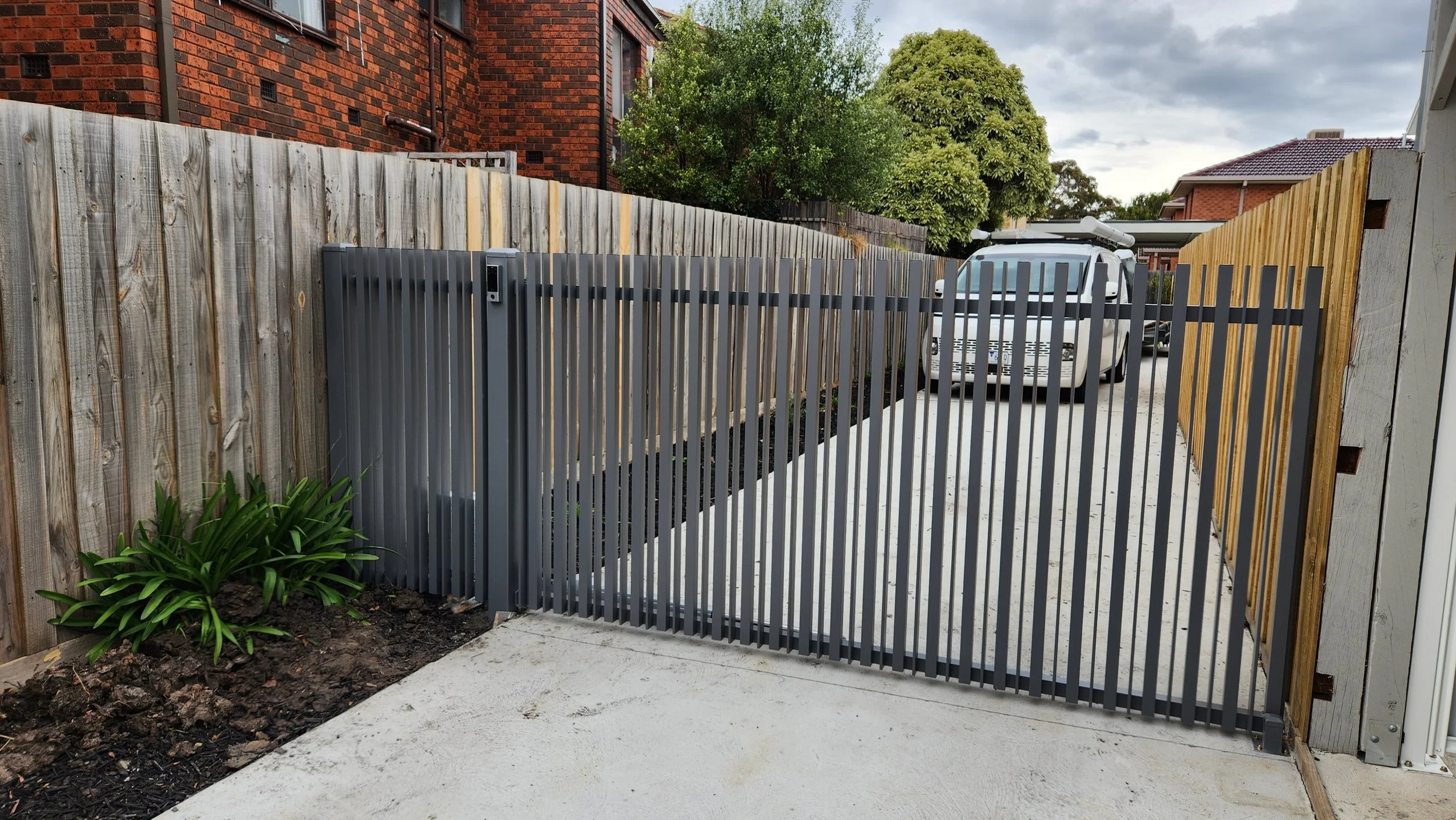 A driveway with a metal gate, a garden bed with green plants on the left, and a white vehicle parked in the background. The surrounding fence has wooden panels and a brick wall.