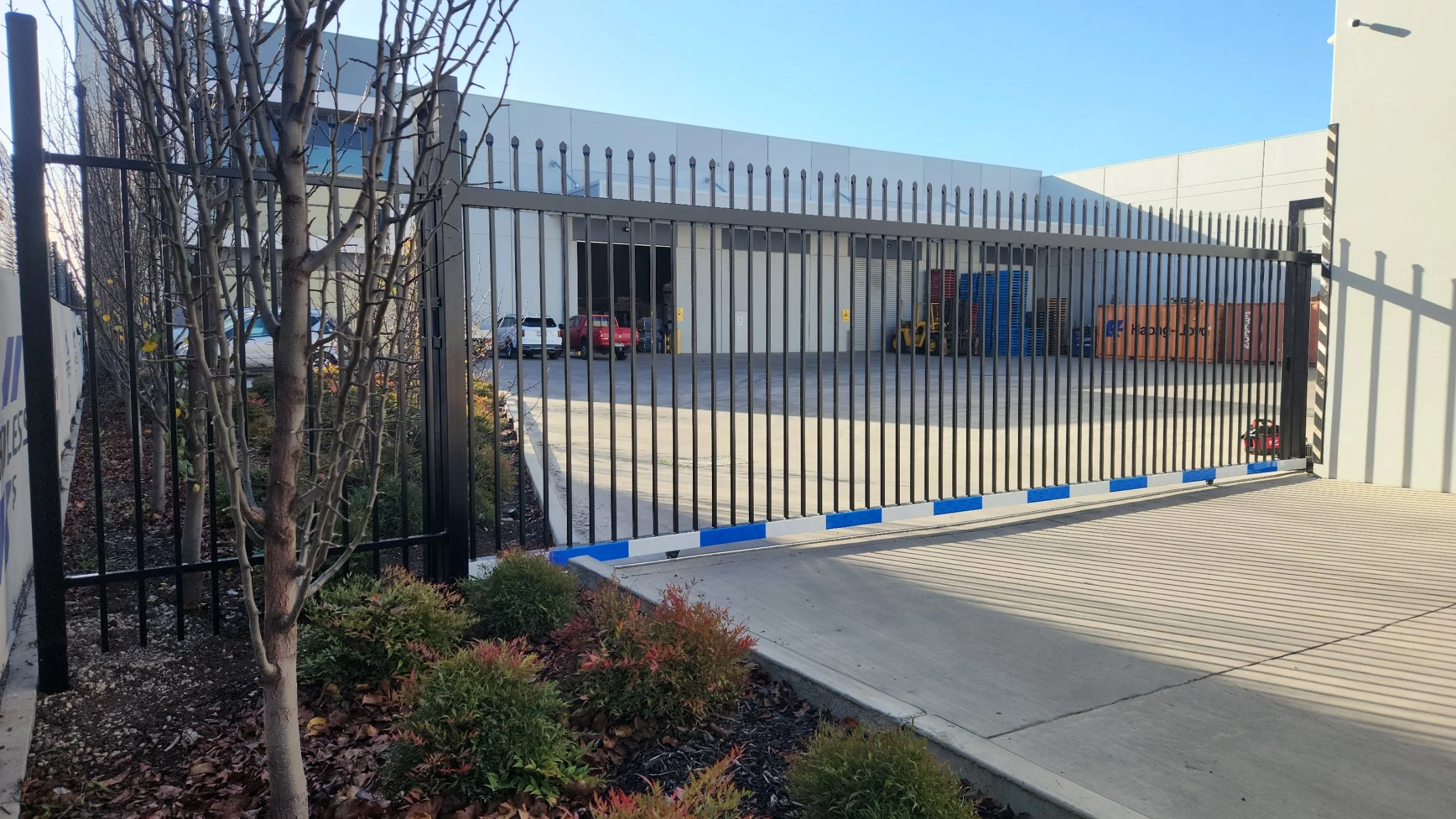 Industrial warehouse with a tall metal gate, parking lot with several parked vehicles, storage containers, and a forklift.