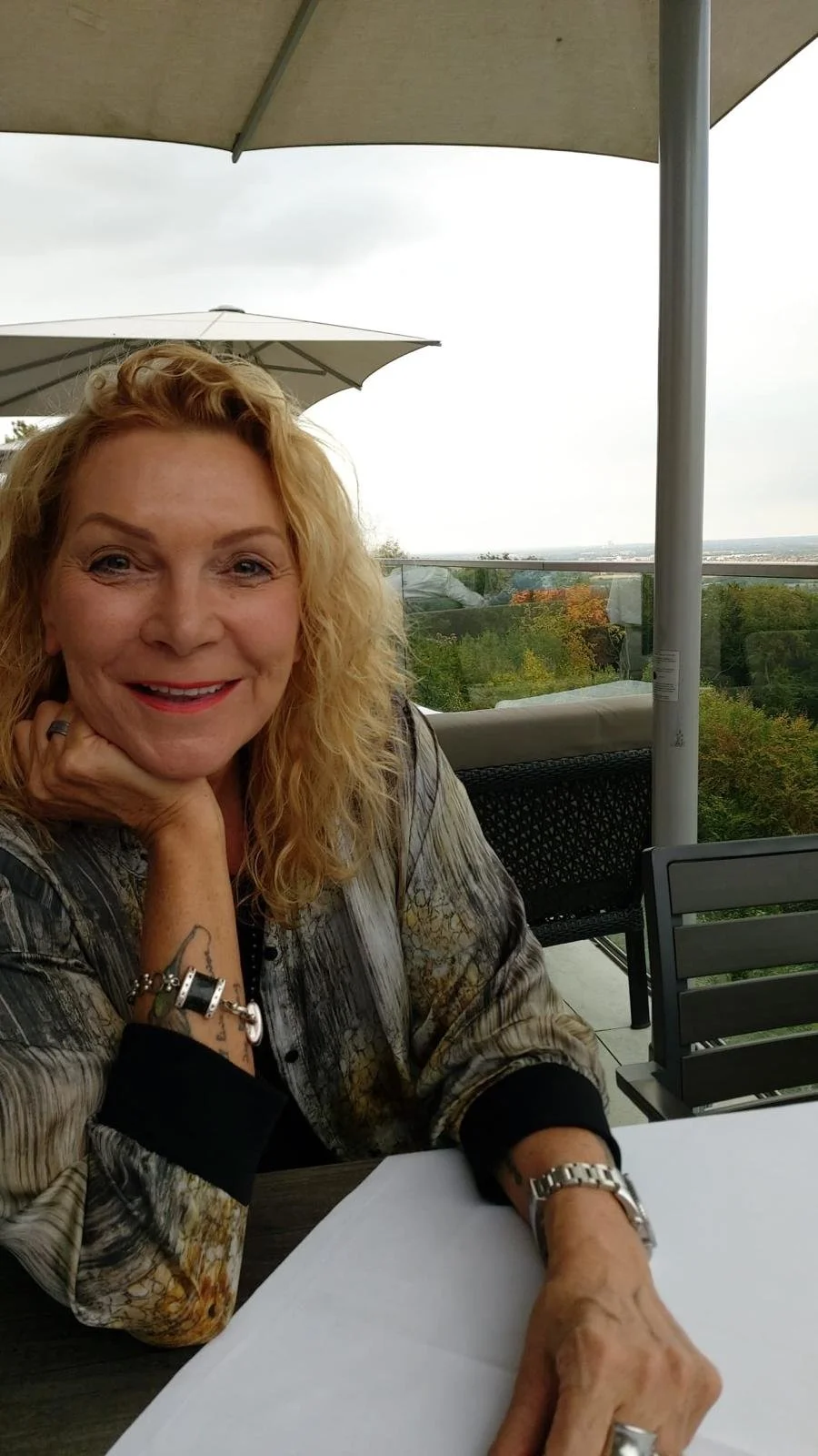 Frau mit blonden Haaren und lächelndem Gesicht sitzt an einem Tisch im Freien, unter einem Sonnenschirm, mit einer Aussicht auf einen bewaldeten Hintergrund.