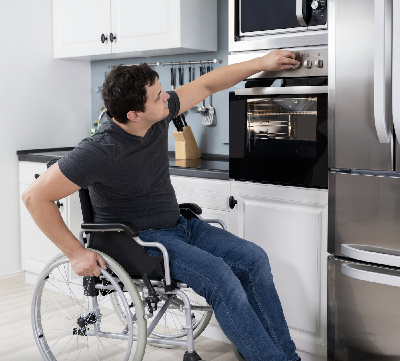 A man in a wheelchair reaching to adjust the control knob of a built-in oven in a kitchen.