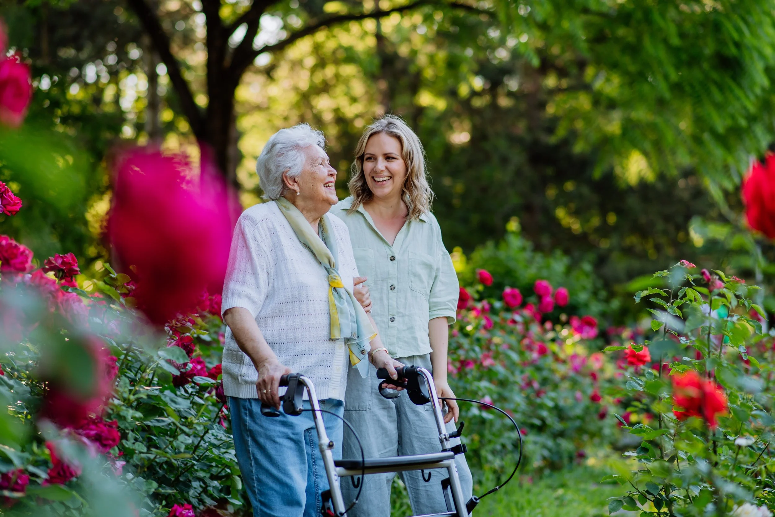 An elderly woman with a walker smiling and talking with a younger woman in a garden surrounded by blooming pink roses and green trees.