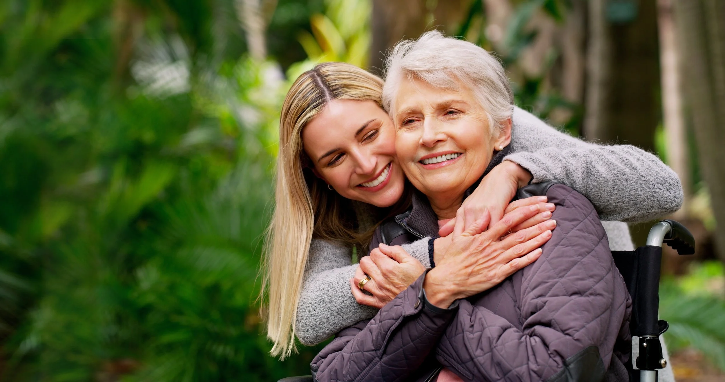 A young woman hugging an elderly woman in a wheelchair outdoors surrounded by greenery.