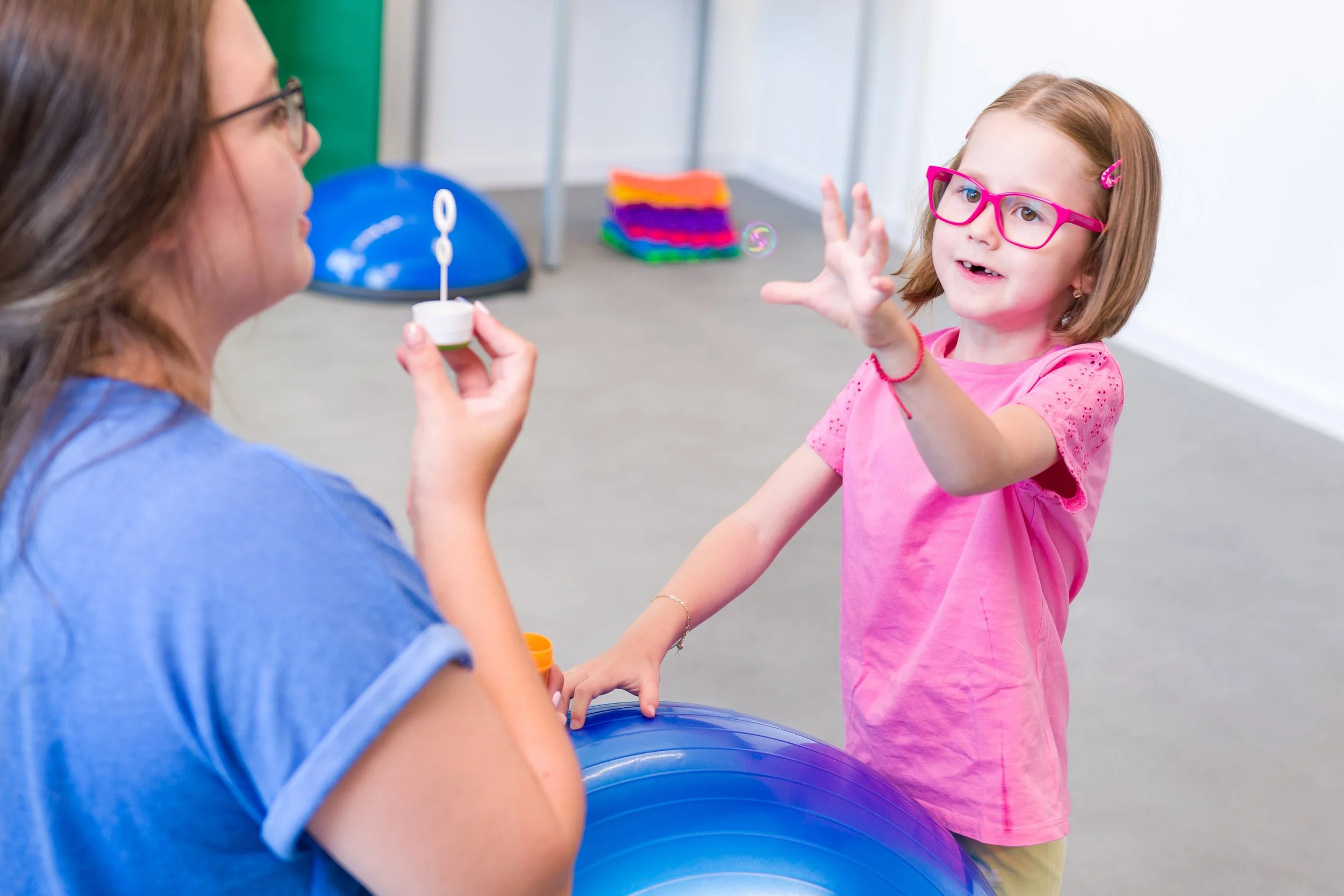 A young girl with glasses and a pink shirt reaches out excitedly while an adult woman blows bubbles towards her in an indoor play area.