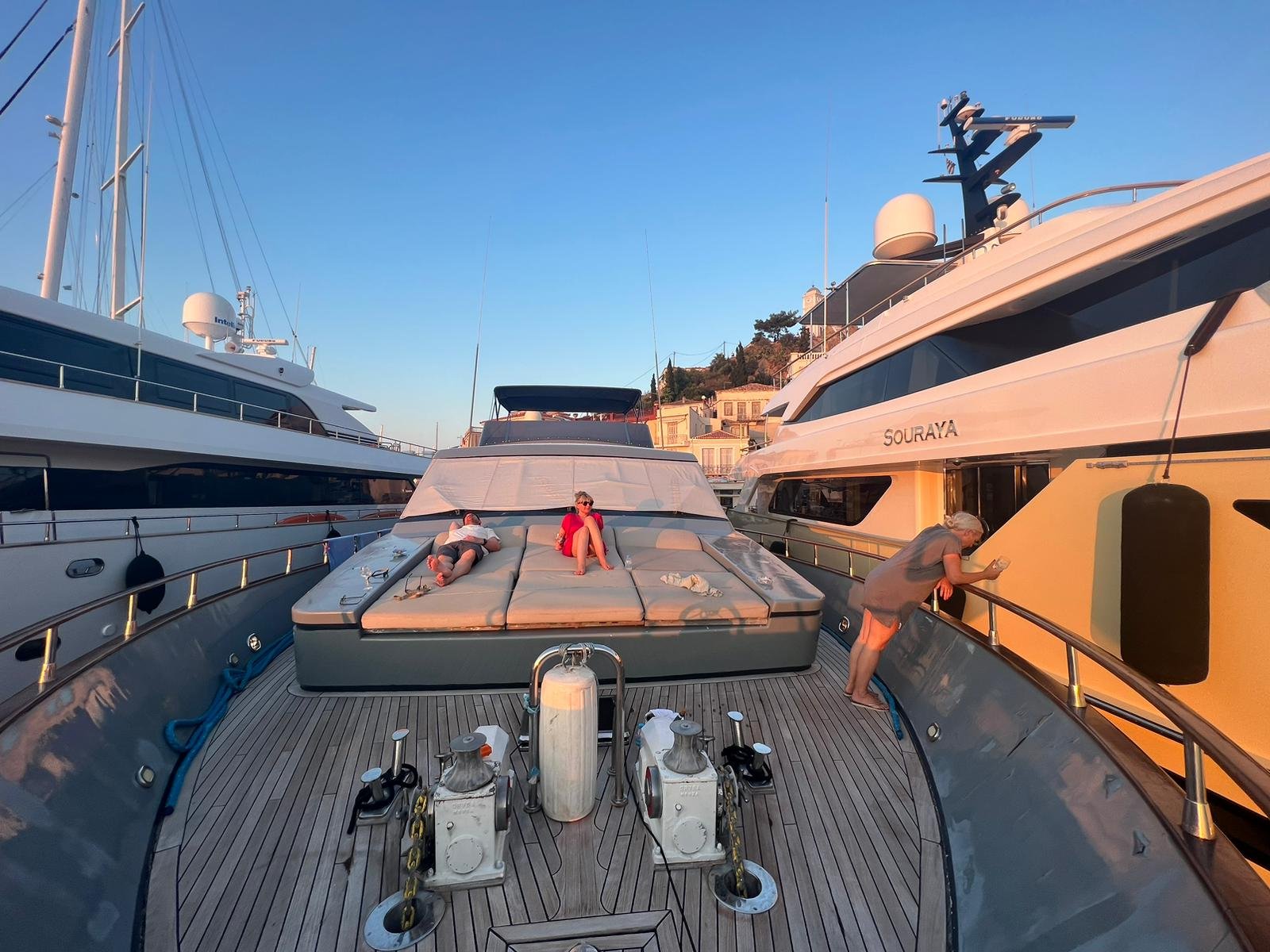 Personen entspannen sich auf dem Deck einer Yacht, während eine weitere Person an der Reling lehnt. Die Boote sind in einem Hafen angedockt, umgeben von weiteren Yachten. Der Himmel ist klar und blau, was auf einen sonnigen Tag hindeutet.