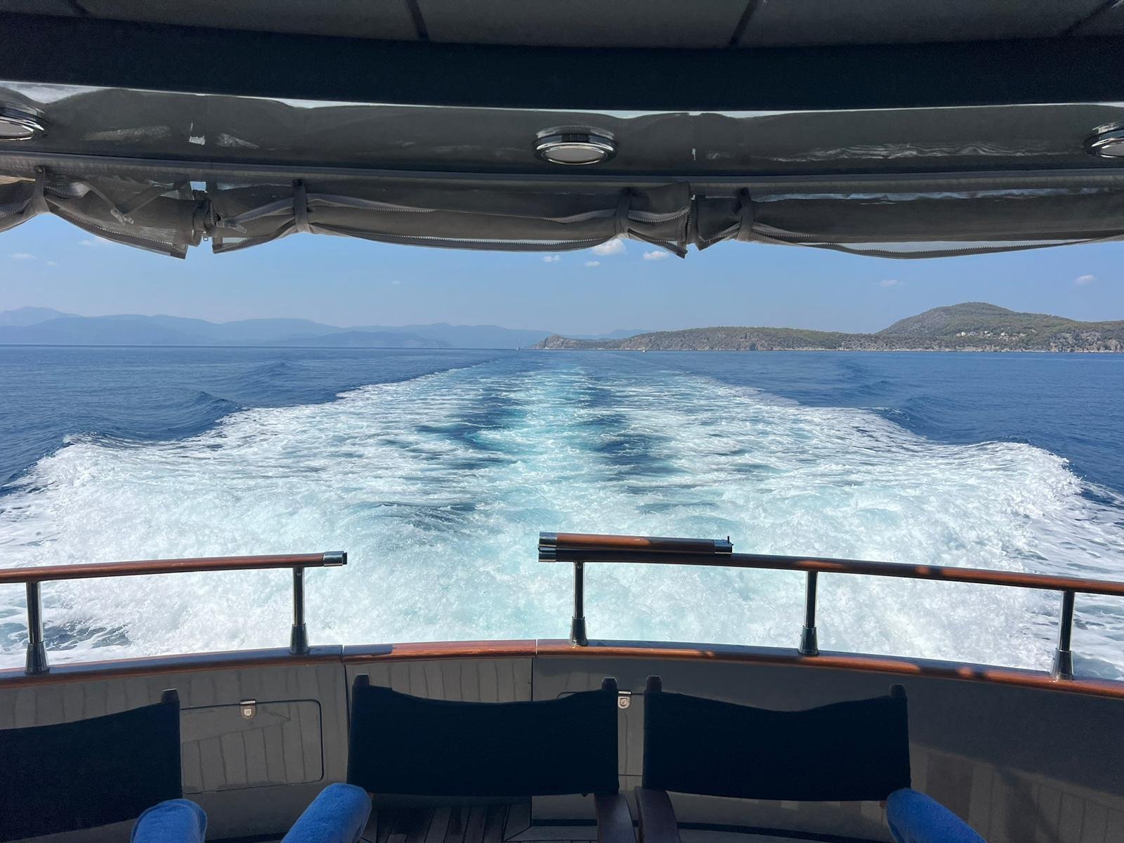 Blick von einem Bootsrücken auf das Meer mit Wasserwellen hinter dem Boot. Im Hintergrund sind Hügel und eine Küste sichtbar. Ein Baldachin hängt über dem Bootsrücken.