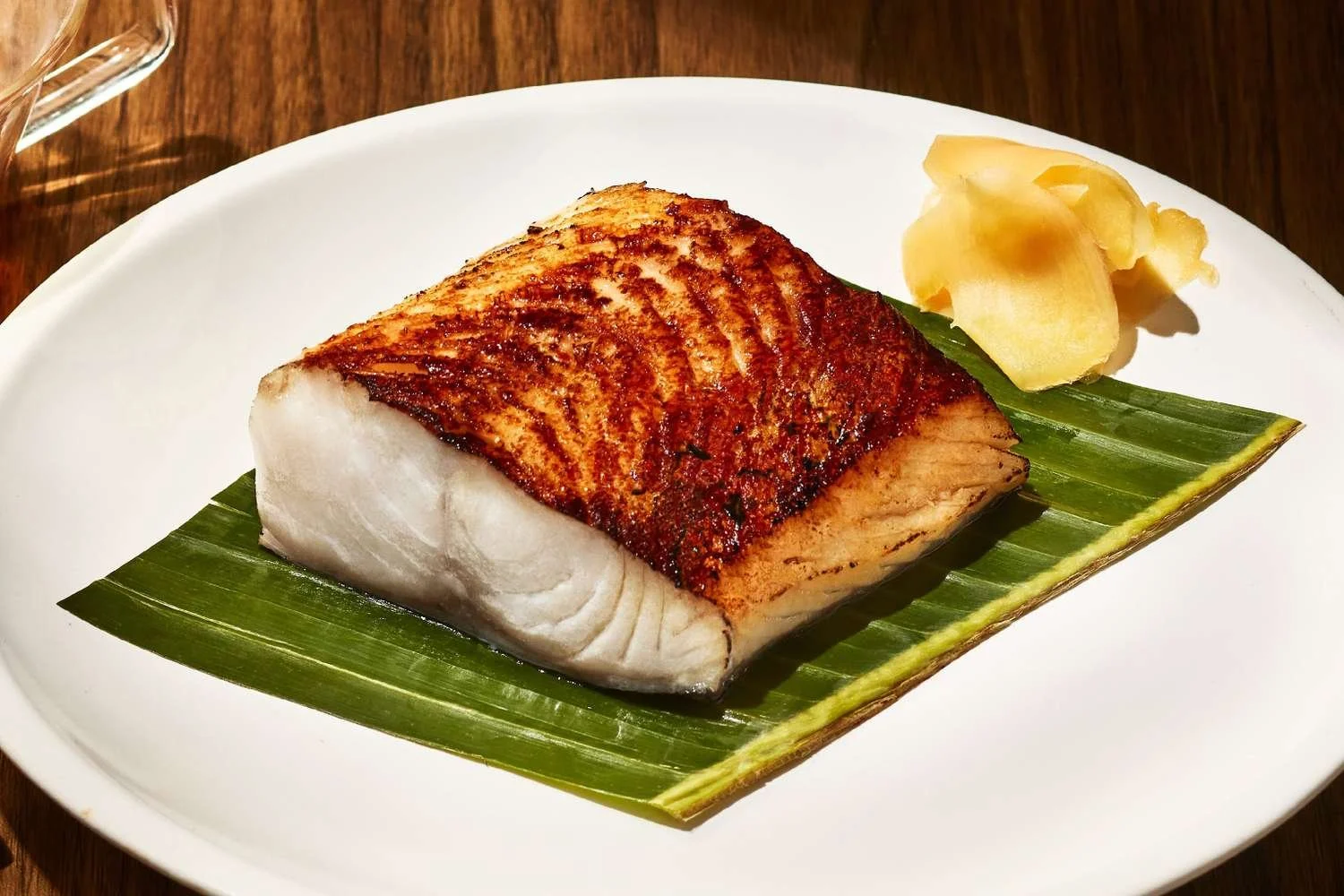 A piece of crispy, grilled salmon with a golden-brown crust, served on a banana leaf on a white plate, accompanied by a portion of sliced ginger.