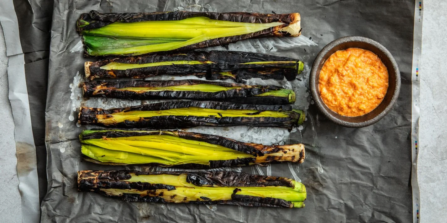 Charred green onions with a bowl of orange sauce on parchment paper.