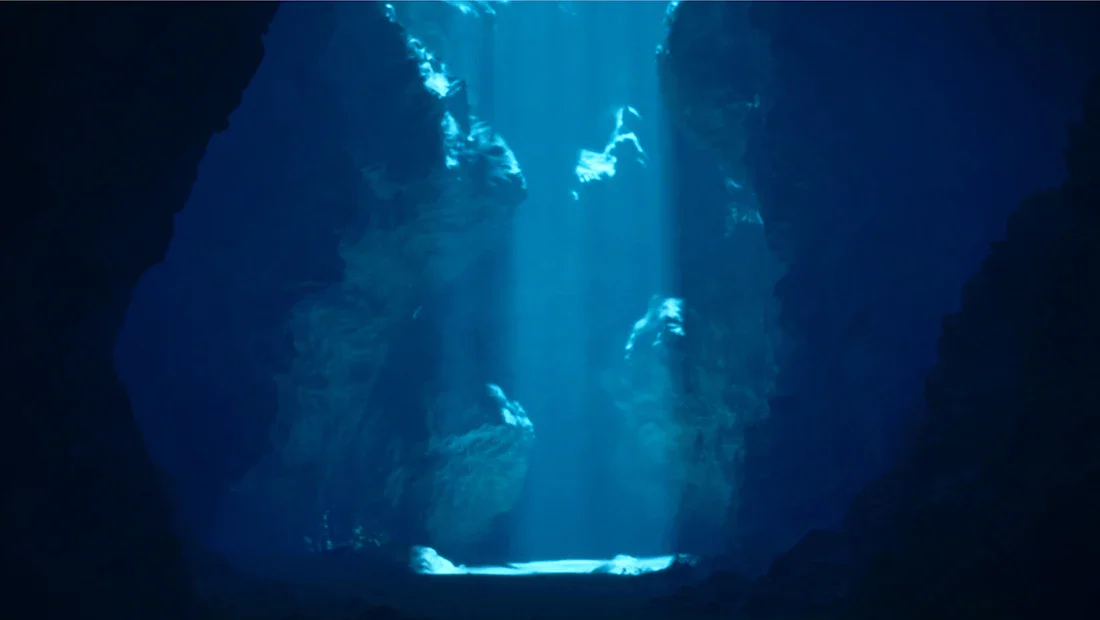 Deep underwater cave with sunlight streaming through opening above, illuminating the water and creating a mystical scene.