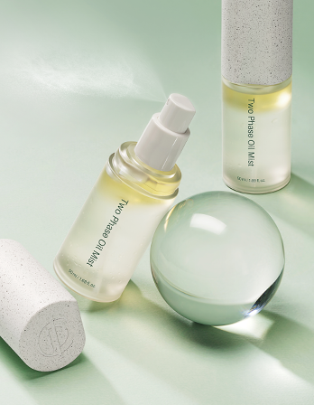 Skincare products including a spray bottle labeled "Two Phases Oil & Mist," a rolled-up white towel, and a glass sphere on a pastel green background.