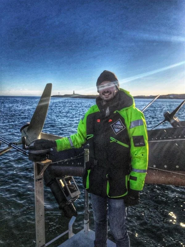 A man wearing a bright green and black jacket, glasses, and a beanie standing on a boat with water and a distant shoreline visible behind him.