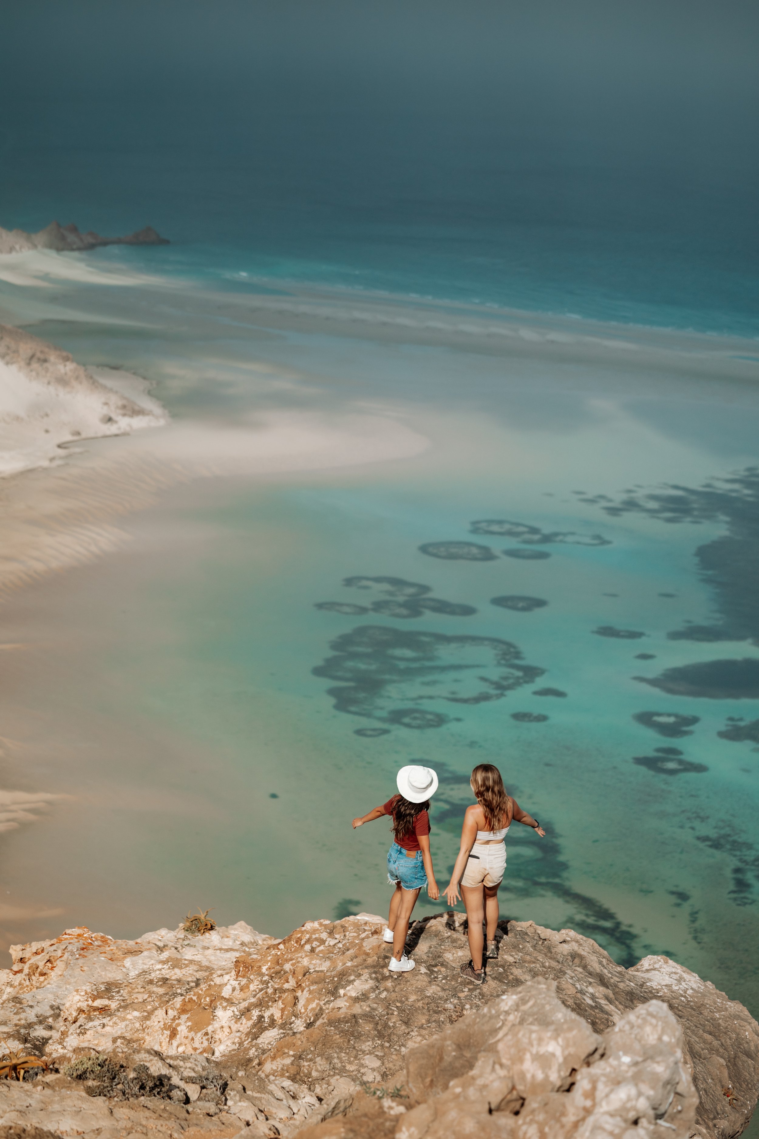 Two people standing on a rocky cliff overlooking a turquoise ocean and sandy shoreline.