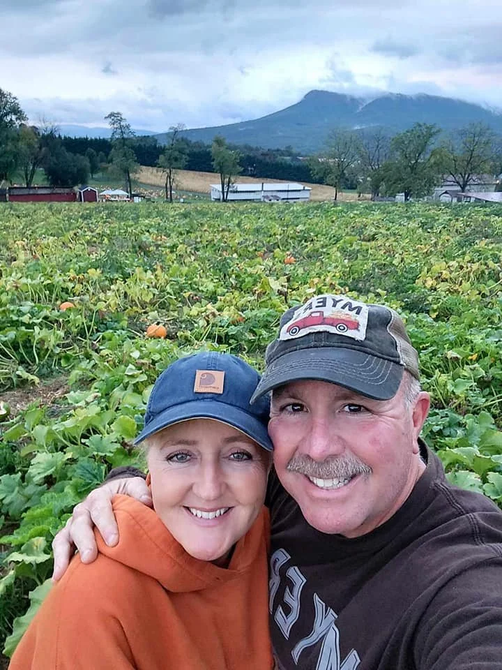 A smiling couple taking a selfie in a pumpkin patch with a mountain in the background.