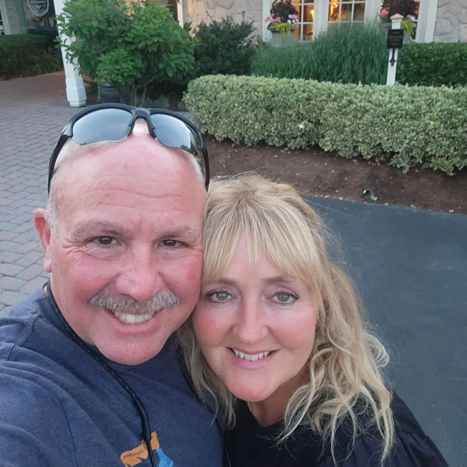 A happy couple taking a selfie outdoors in front of a house with well-maintained bushes and flowers. The man has a mustache, glasses on his head, and is wearing a dark shirt. The woman has blonde hair and is smiling.