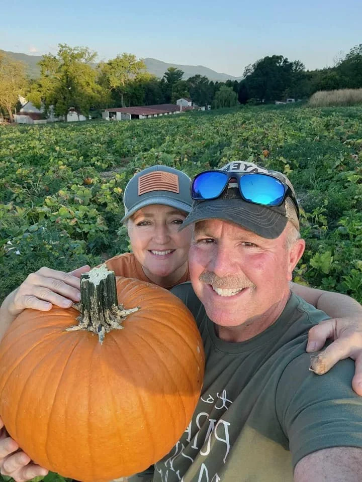 A smiling couple taking a selfie in a pumpkin patch, holding a large pumpkin, with green foliage and distant mountains in the background.