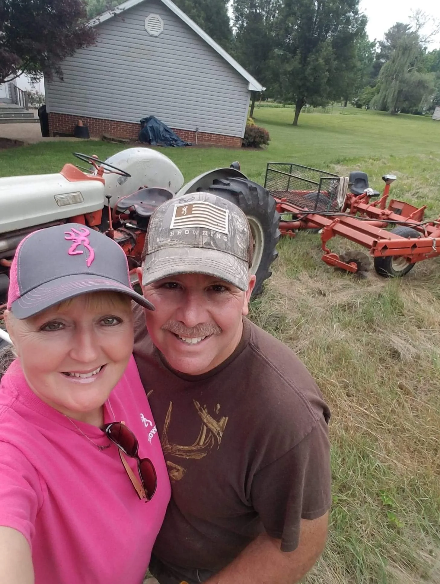 A smiling woman and man taking a selfie outdoors on a farm or rural property with overlapping farming equipment, including a tractor, a mower, and other machinery, in the background.