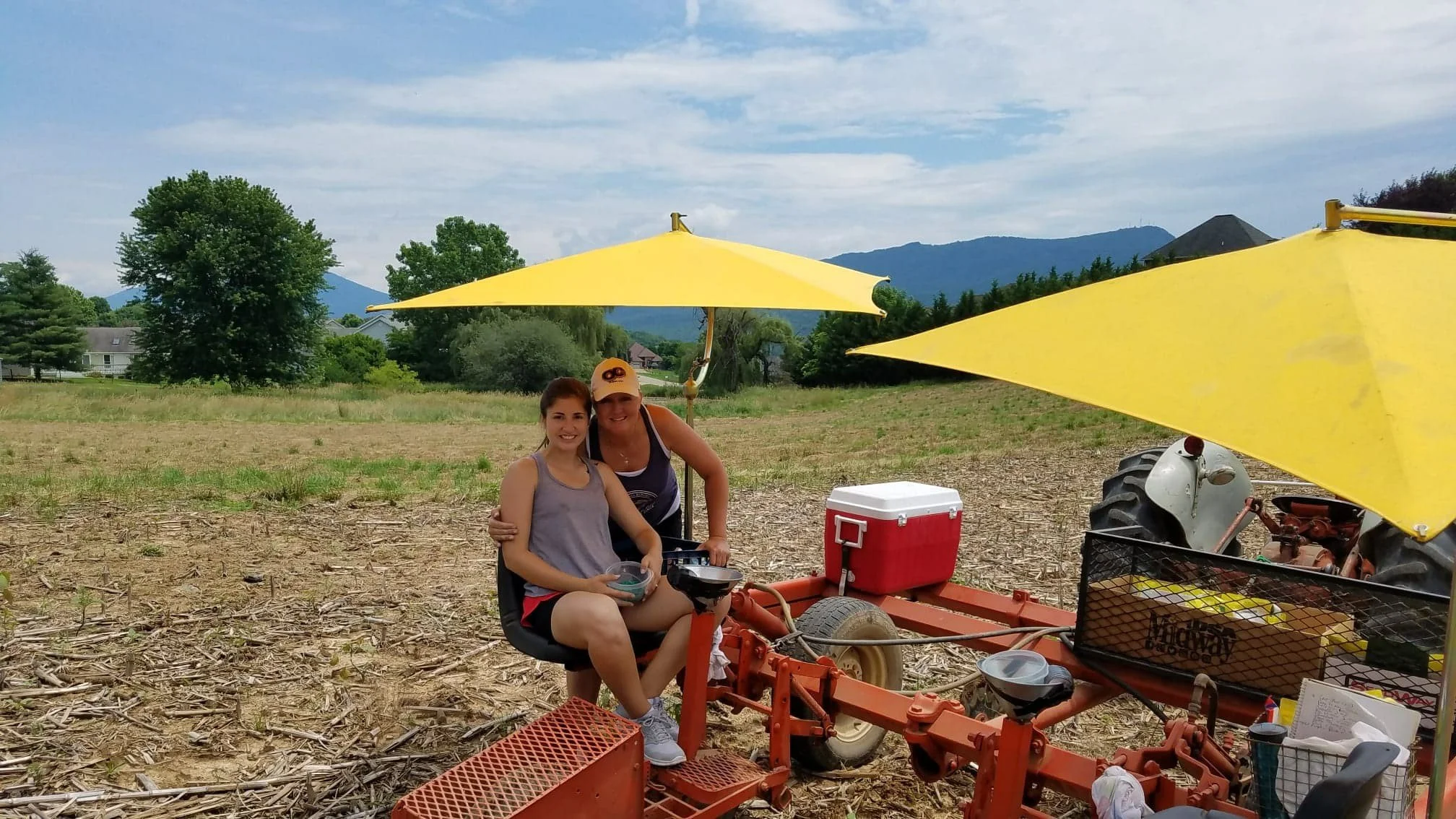Two women smiling and sitting on an orange farm tractor in a field with trees and mountains in the background. Yellow umbrellas providing shade, a red cooler, and various tools and containers are also visible.