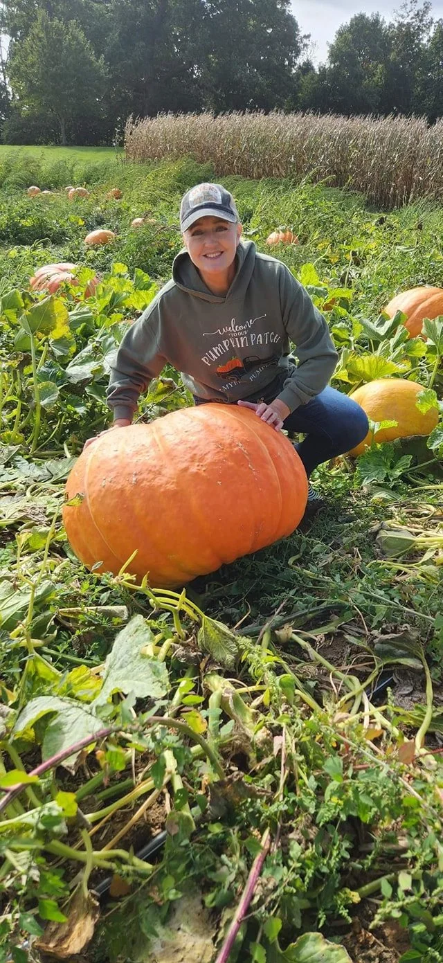 A smiling woman in a gray hoodie and baseball cap crouching next to a large pumpkin in a pumpkin patch, with more pumpkins and a field of tall plants in the background.