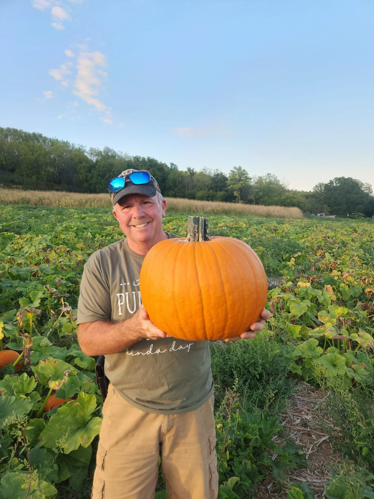 A man smiling and holding a large orange pumpkin in a pumpkin field on a clear day.