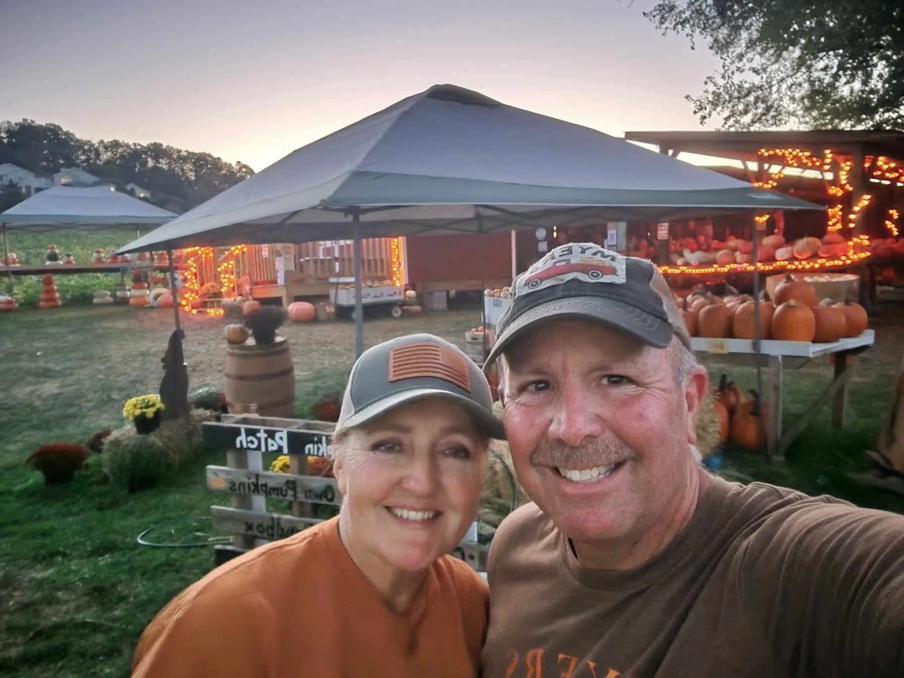 Smiling man and woman taking selfie at a pumpkin patch with pumpkins displayed on tables, string lights, and tents in the background during sunset.