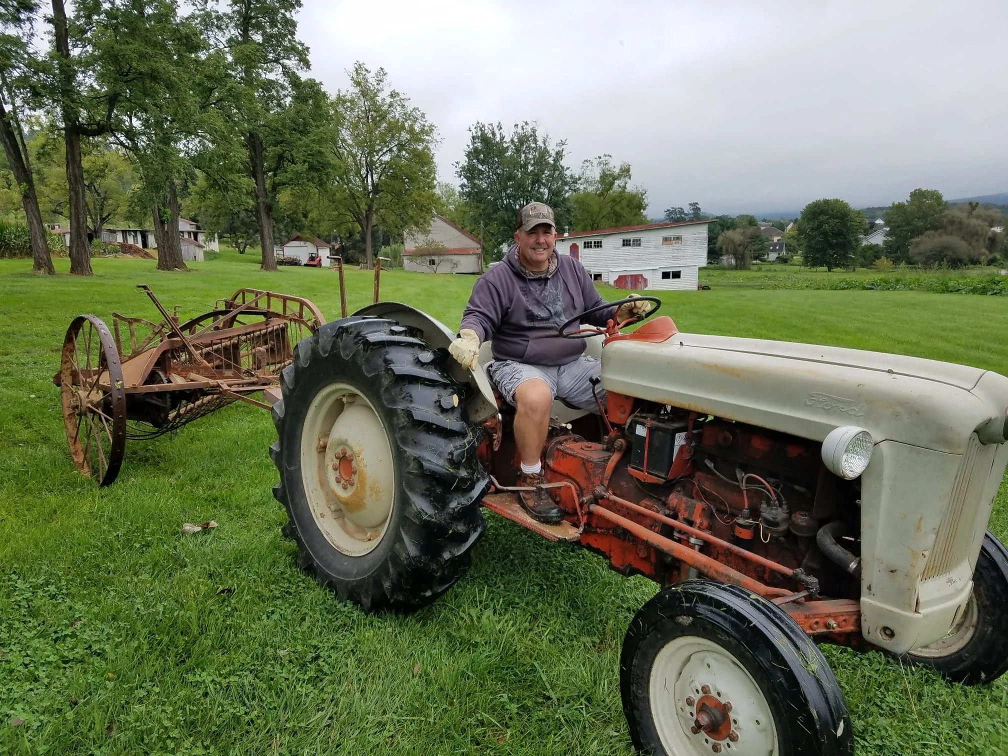 A man sitting on an old Ford tractor on a grassy field with trees and buildings in the background.