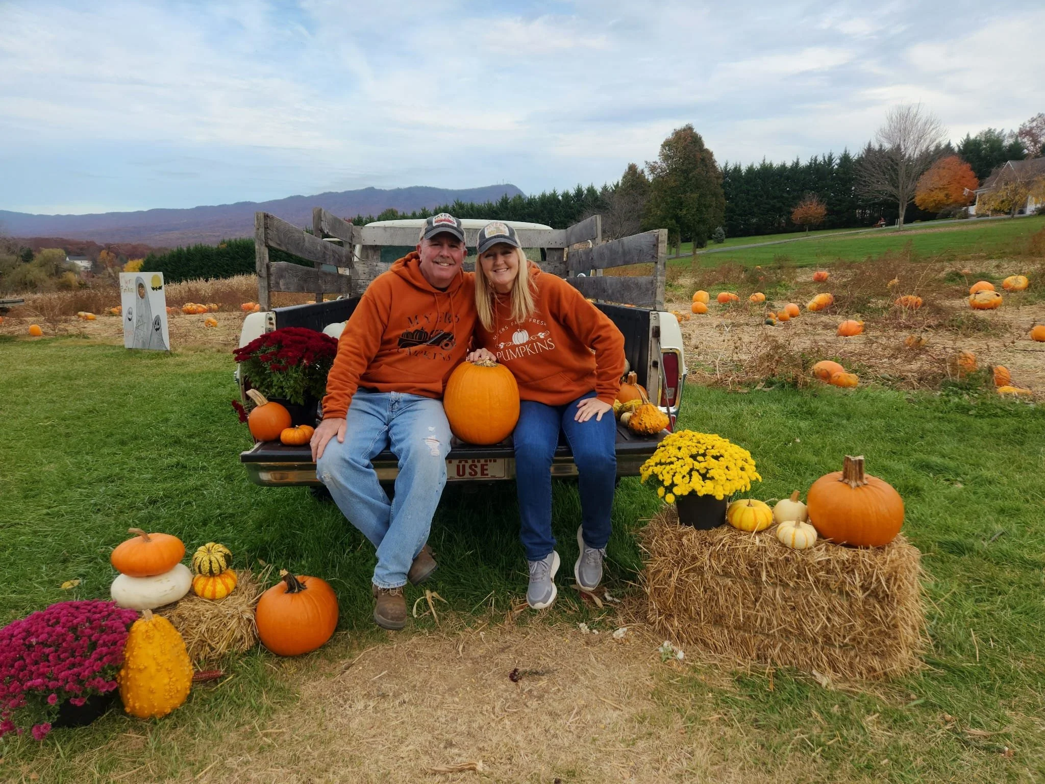A smiling couple sitting on the tailgate of a truck surrounded by pumpkins, gourds, and colorful fall flowers at a pumpkin patch.