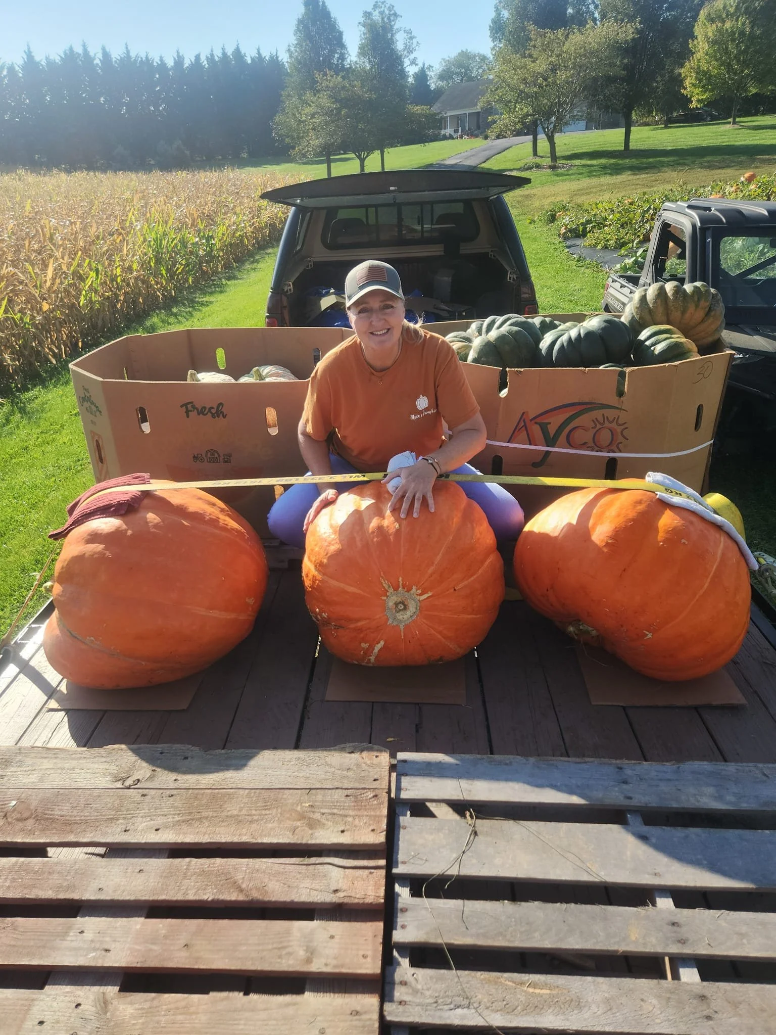 A woman sitting on a flatbed truck filled with large pumpkins and squash, in a rural farm setting with fields and trees in the background.