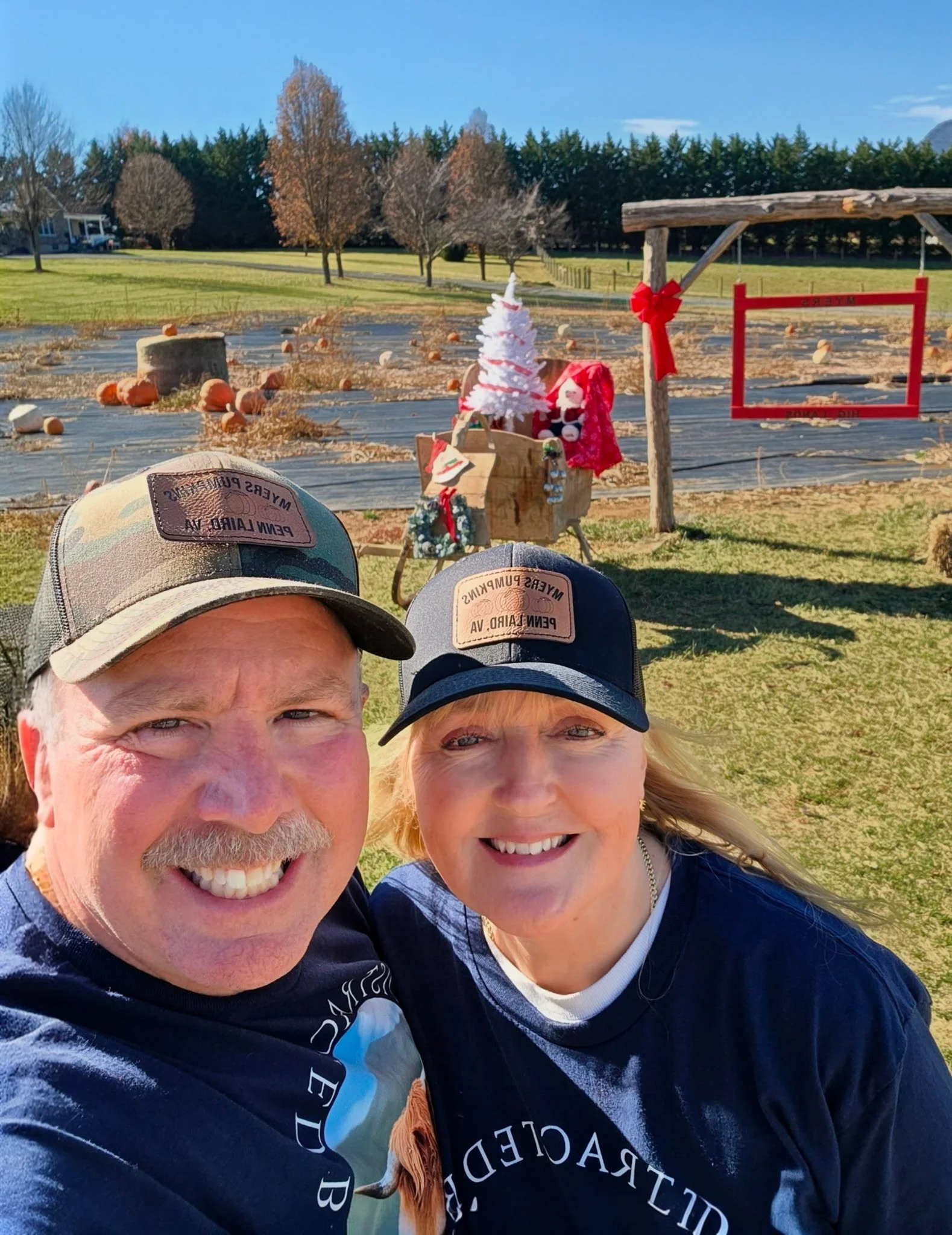 A smiling couple taking a selfie outdoors near a festive display with a small decorated Christmas tree, plush toys, and a red ribbon. The background features a rural scene with pumpkins, trees, and a clear blue sky.