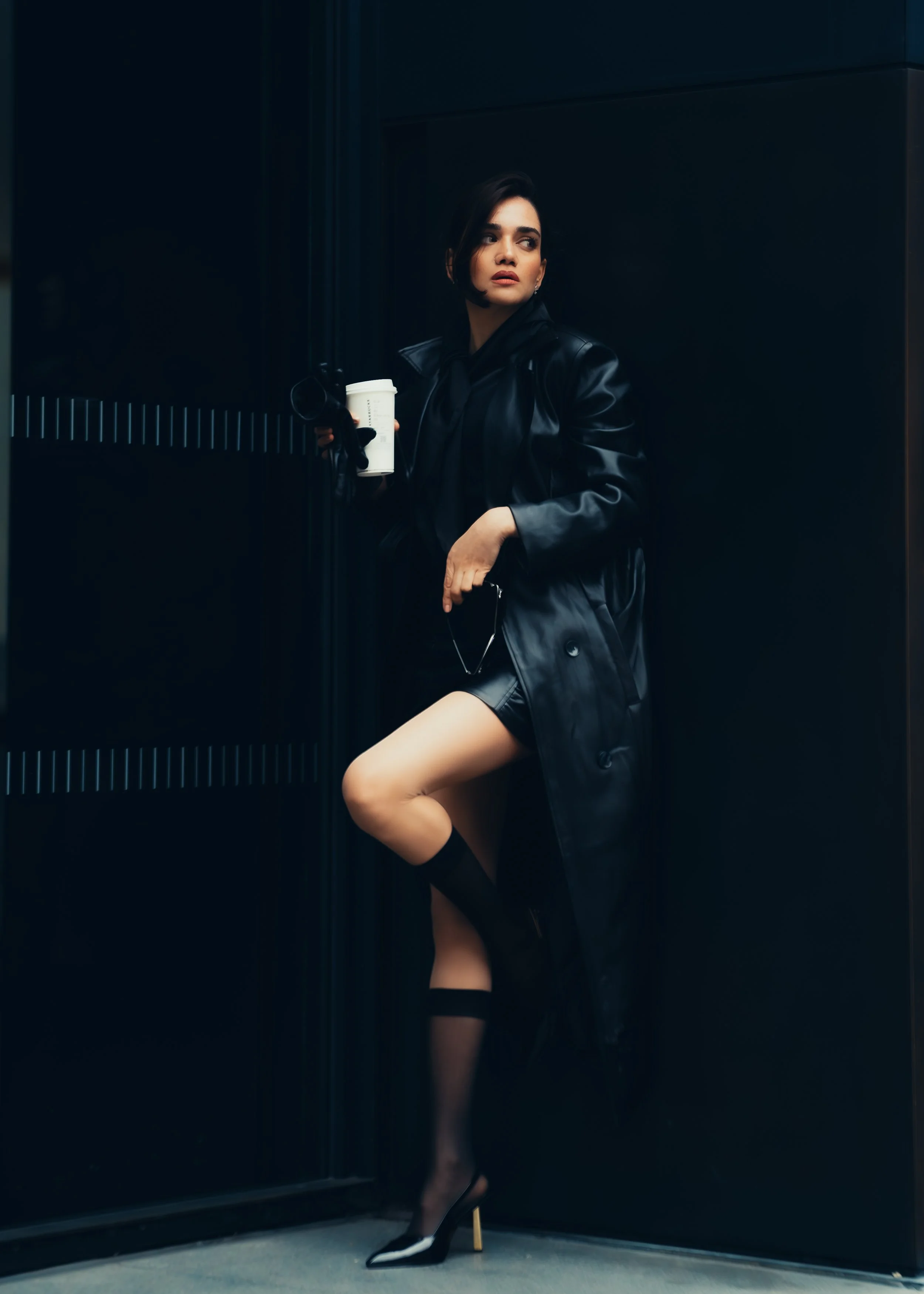 A woman in a black leather trench coat, black dress, and black high heels standing in a dark corner, holding a coffee cup, with her left leg lifted and leaning against the wall.