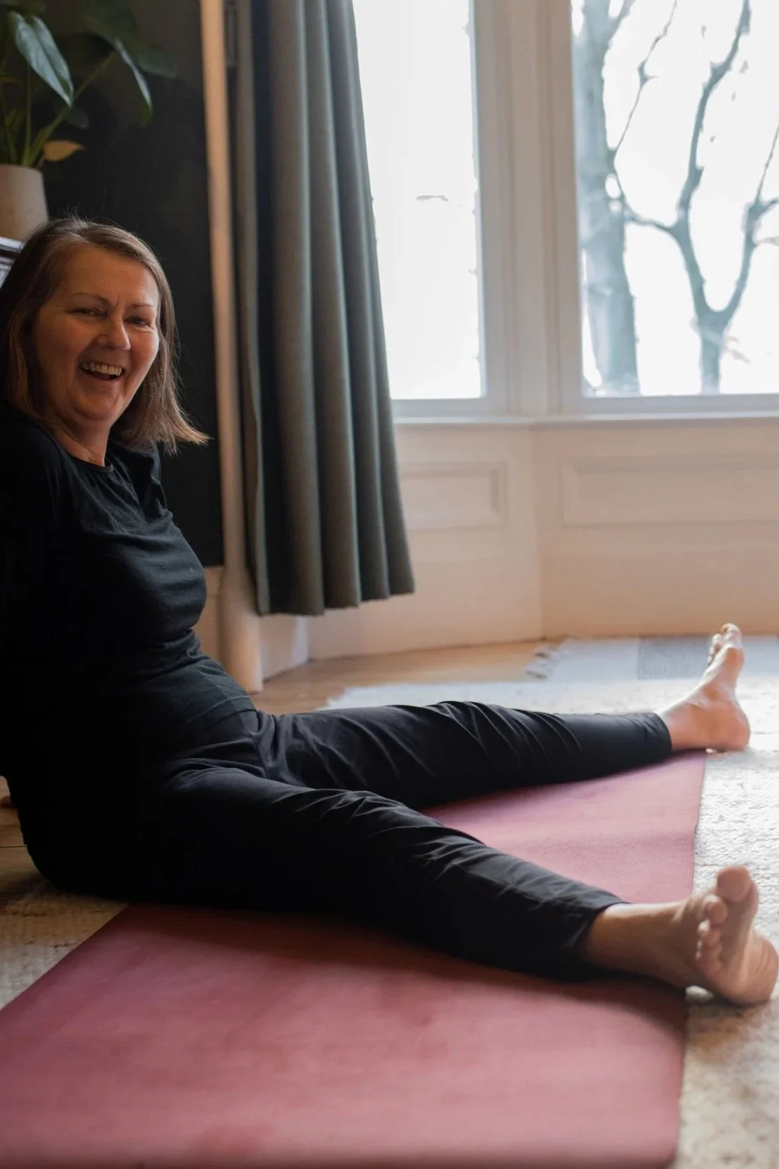 A smiling woman practicing seated straddle stretch on a red yoga mat in a room demonstrating Mat Pilates