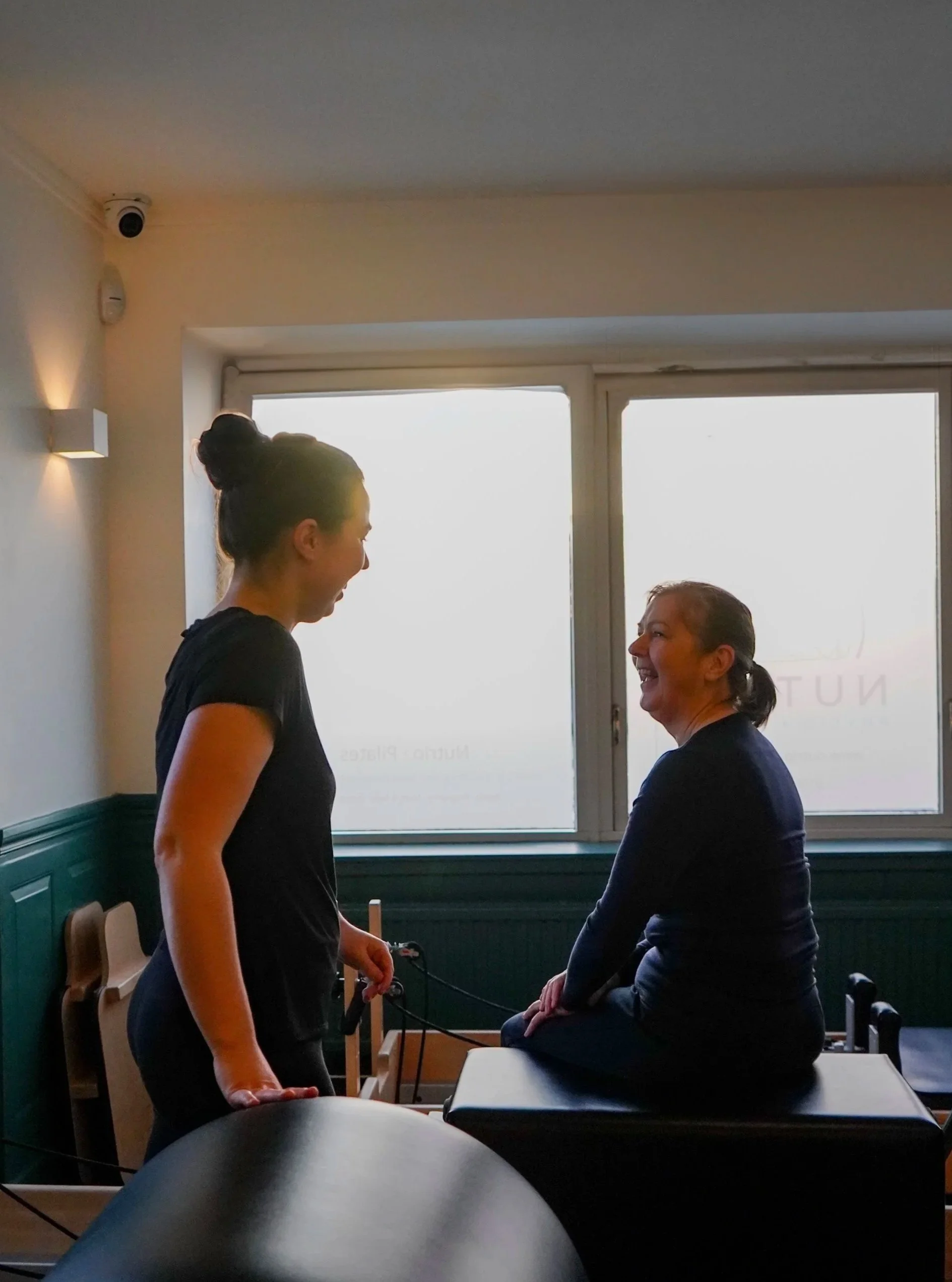 Two women in a pilates studio, smiling and engaging in conversation, with one sitting on a reformer  and the other standing.