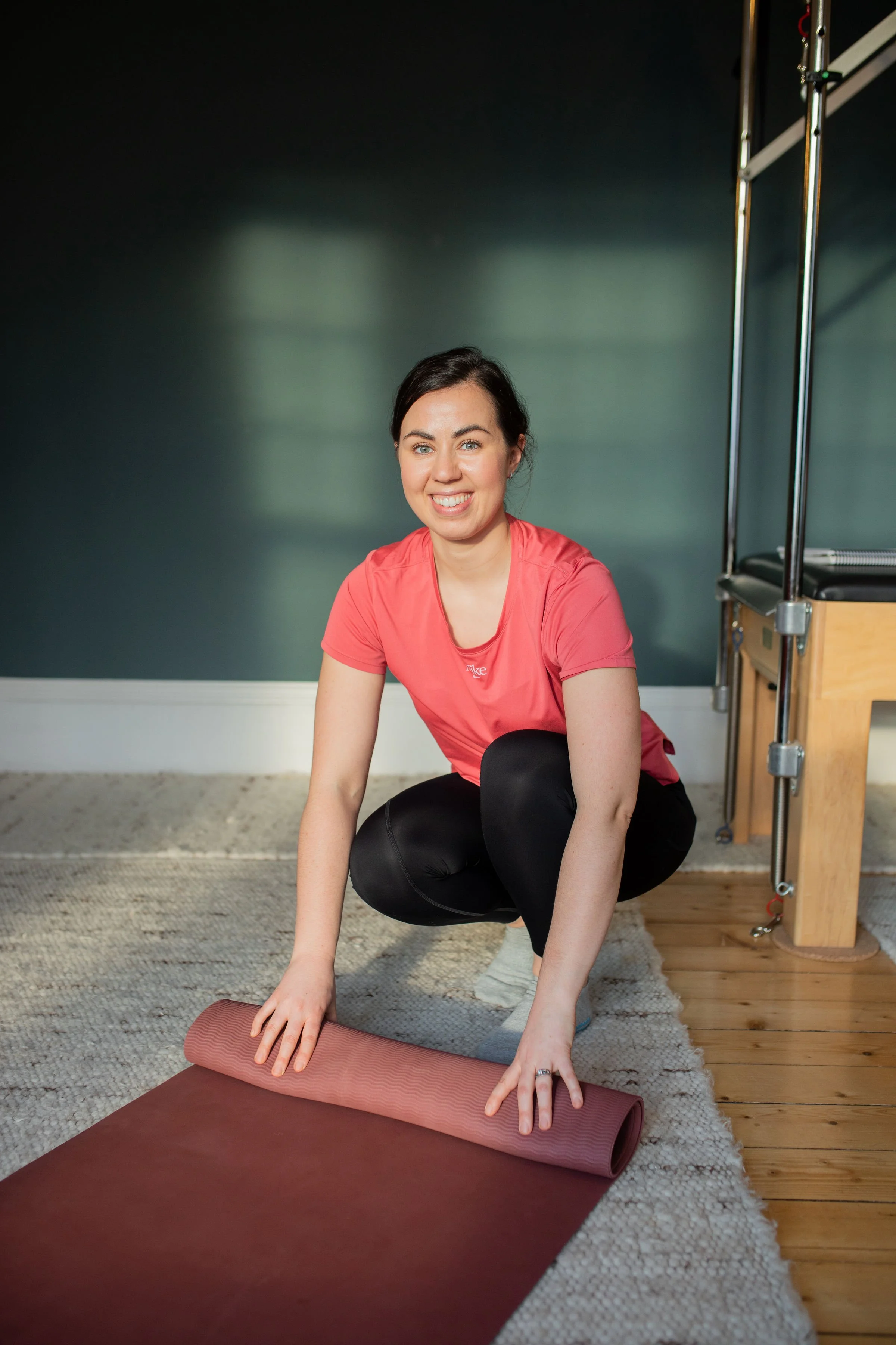 A physiotherapist with dark hair smiling while kneeling on a pilates mat, preparing to do yoga, pilates or stretch.