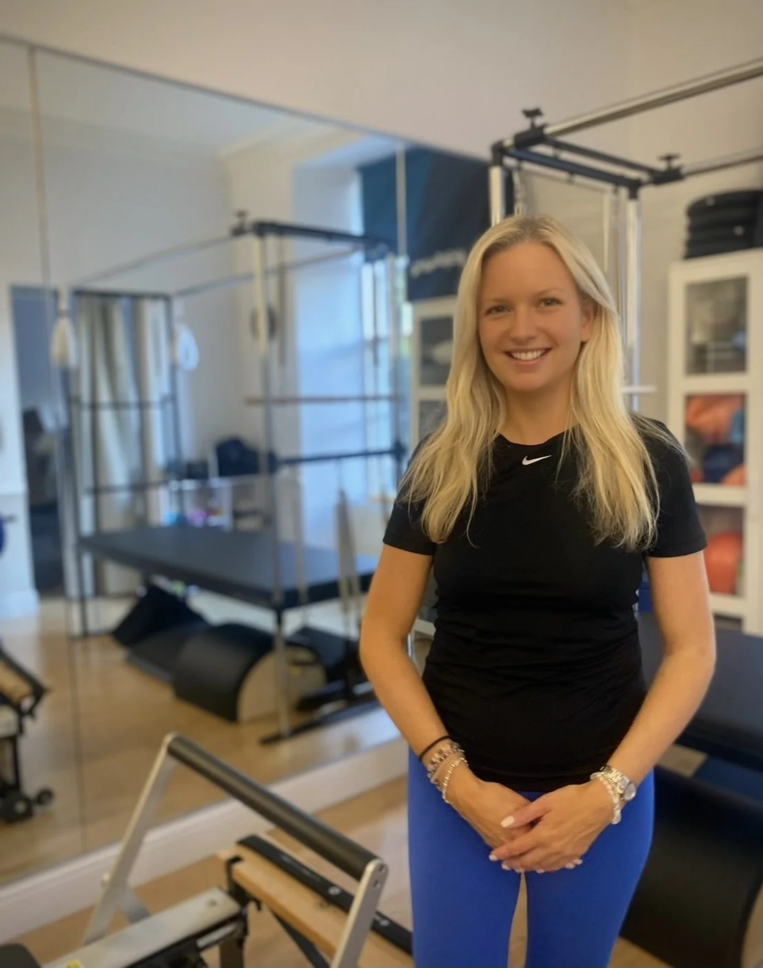 A woman in athletic attire standing in a pilates studio with workout equipment in the background.
