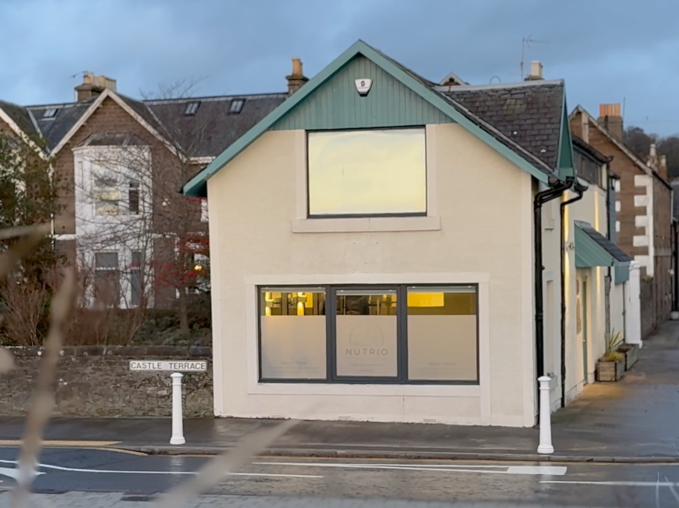 Physiotherapy clinic with a white facade and green roof, with large windows on both floors, situated on Broughty ferry street corner with a pedestrian sidewalk, and a street sign labeled 'Castle Terrace' in front.