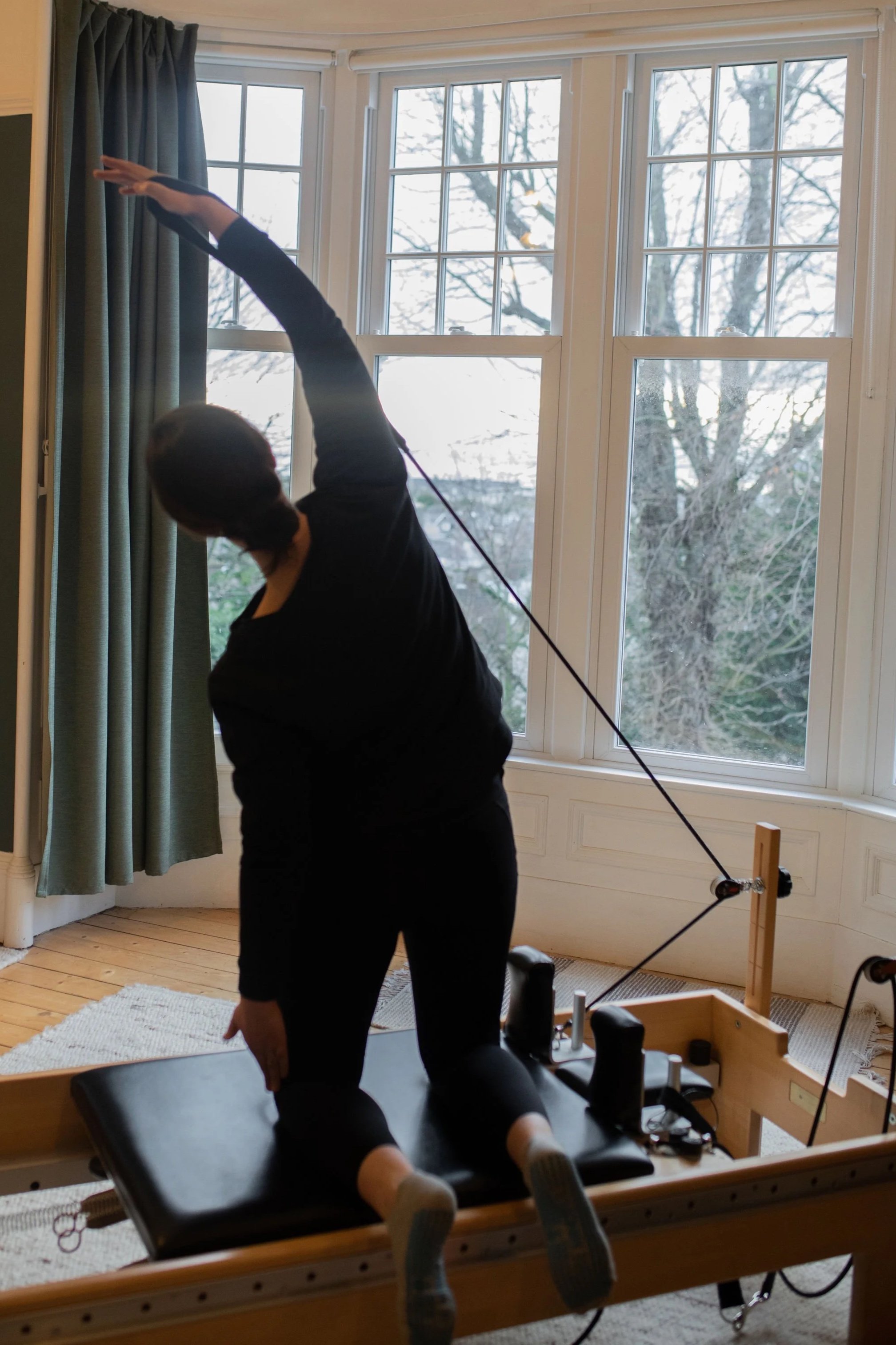 A woman is performing a stretching exercise on Pilates equipment in a room with large windows and a view of trees outside.