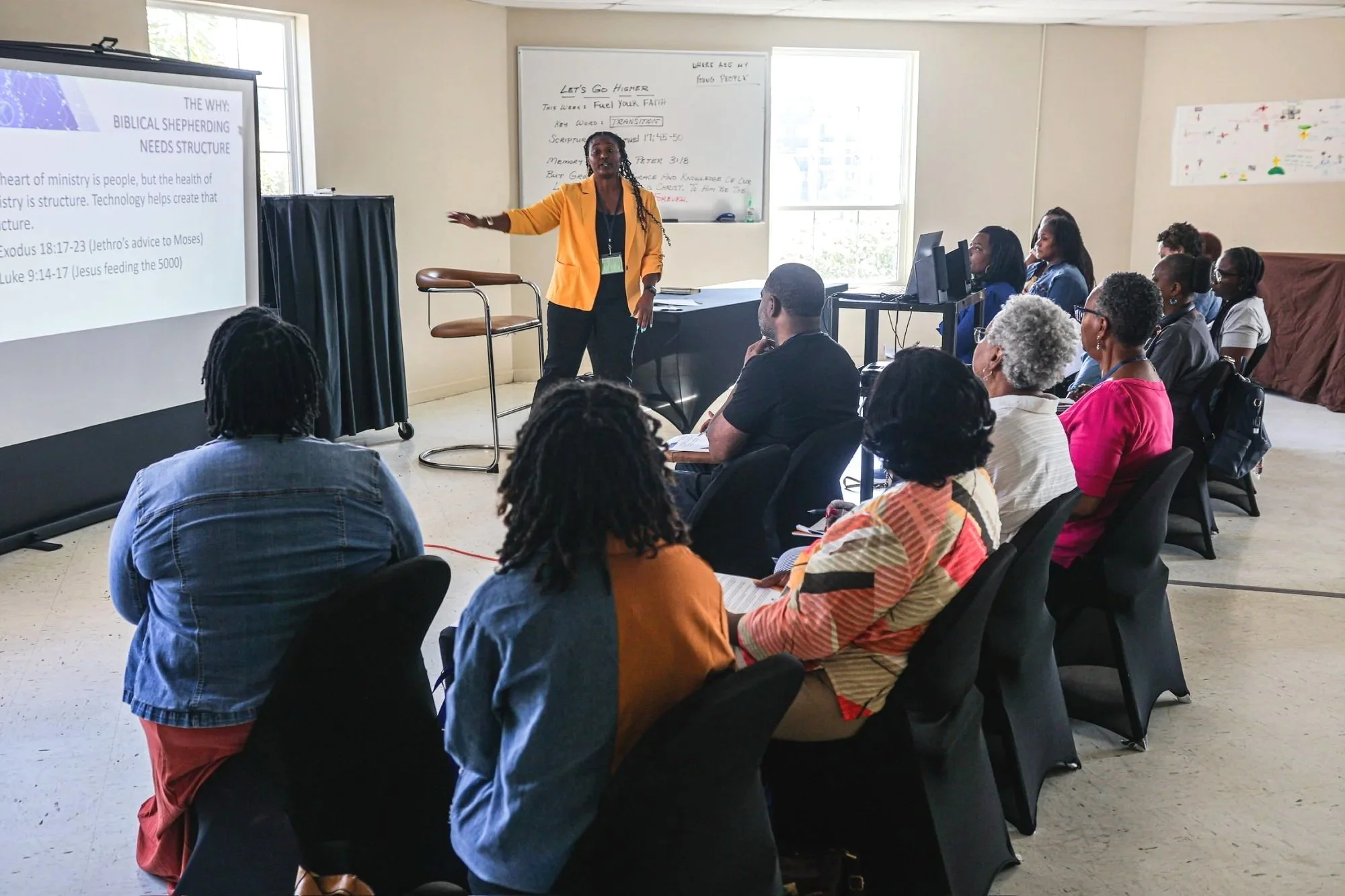A woman in a yellow blazer gives a presentation to an audience in a conference room. The presentation slide is visible on a screen, and the audience is seated and listening attentively.