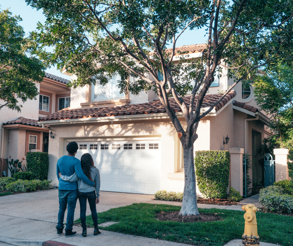 A couple stands together on a suburban street at sunset, facing a two-story house with a tiled roof and a large tree in the front yard.