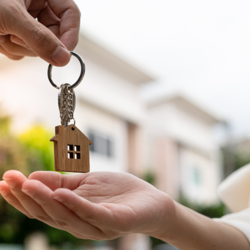 Person handing over a house-shaped keychain to another person outdoors.