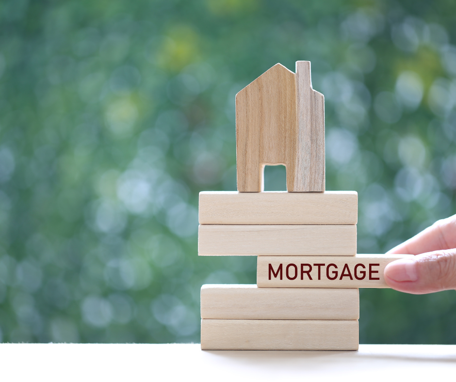 A small wooden house model balanced on wooden blocks labeled 'MORTGAGE', with a person's hand holding the right side, set against a blurred green outdoor background.