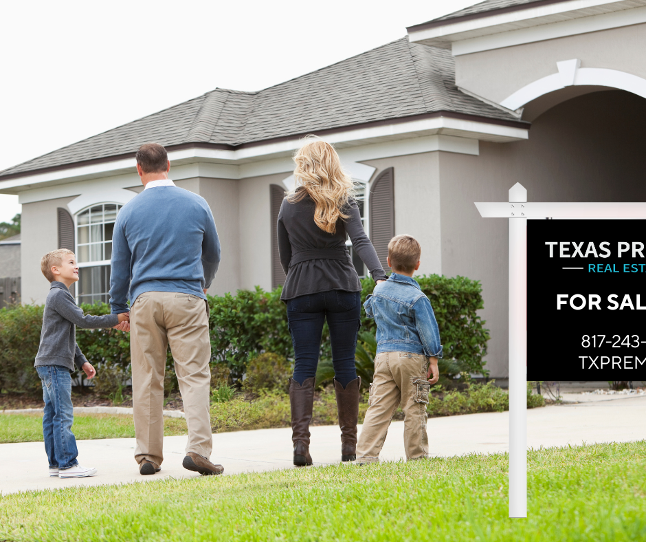 Family of four, two adults and two children, standing outside a house with a 'For Sale' sign in front of it.